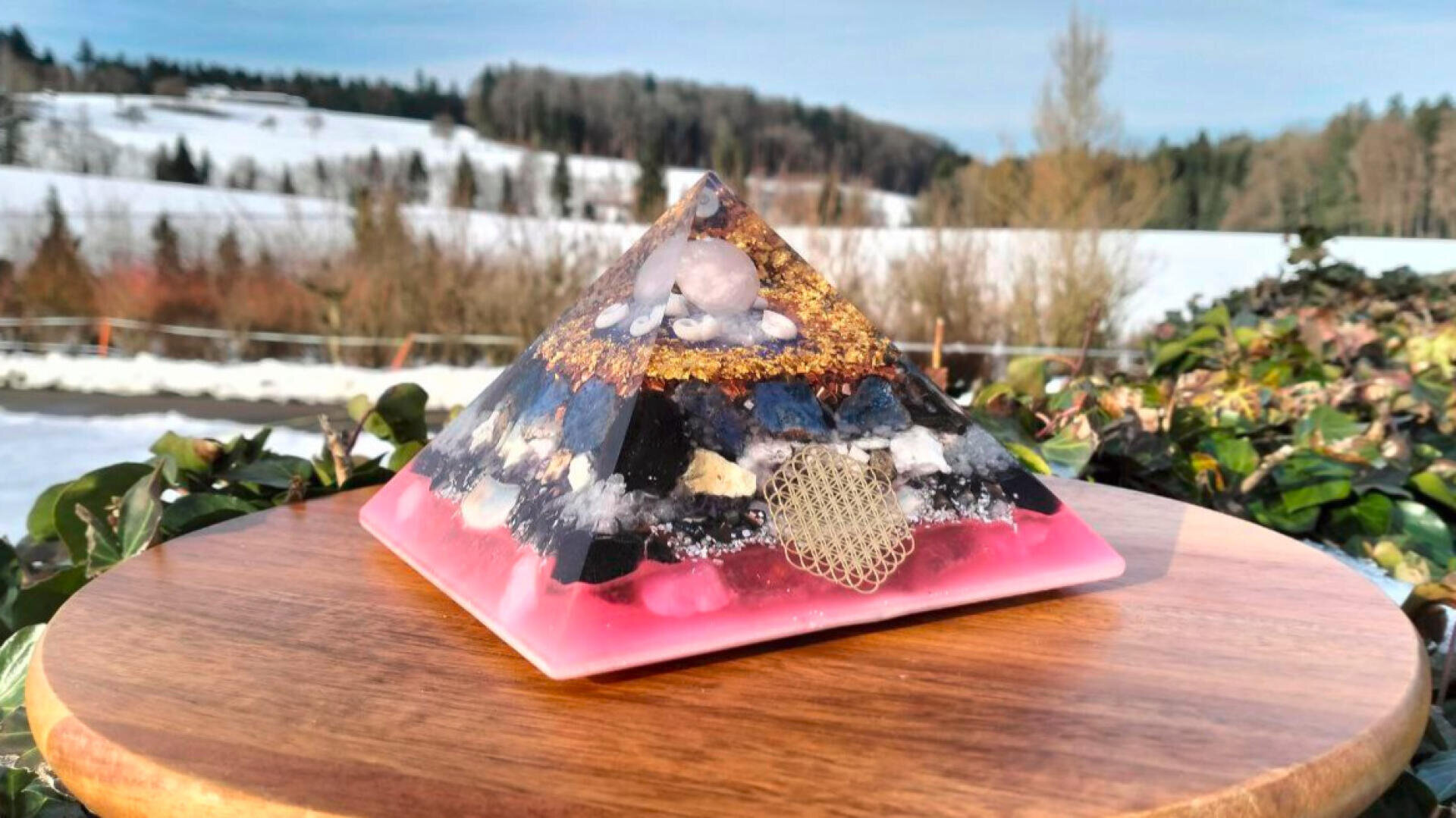 Eine farbenfrohe Harzpyramide mit Steinen und einem Herz aus Golddraht sitzt auf einer hölzernen Oberfläche im Freien, mit verschneiten Feldern, Bäumen und einem blauen Himmel im Hintergrund.
