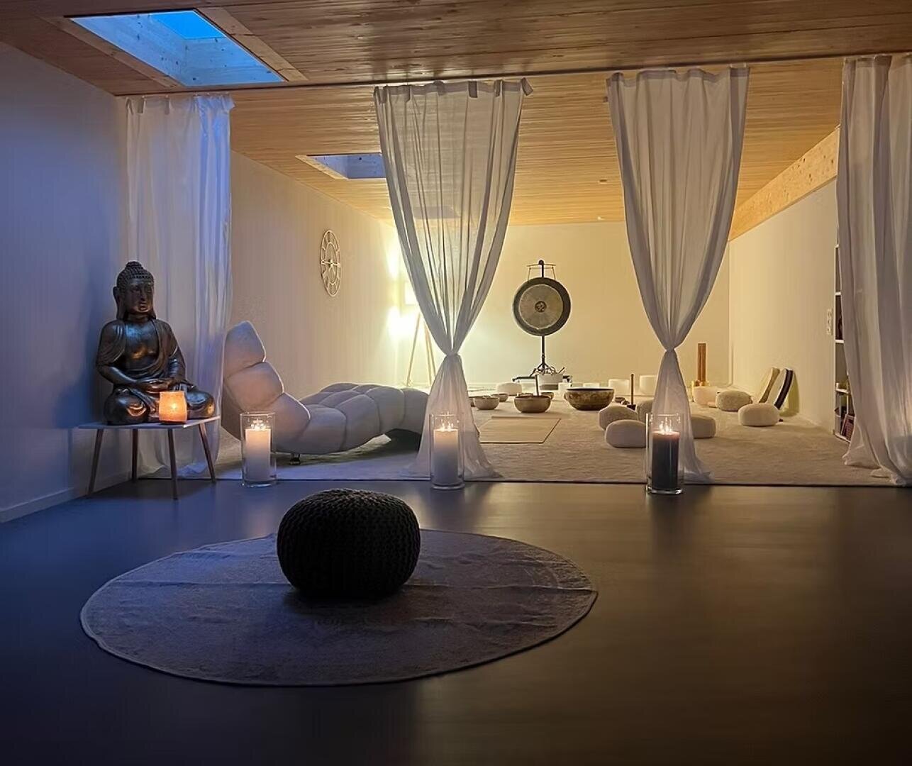Ein friedlicher Meditationsraum mit sanfter Beleuchtung, weißen Vorhängen, Kerzen, einer Buddha-Statue, gepolsterten Sitzgelegenheiten, einem Gong und dekorativen Schalen, die eine beruhigende und gelassene Atmosphäre schaffen.