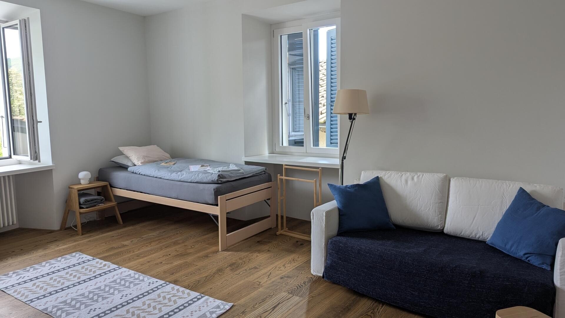 Ein helles, minimalistisches Zimmer mit einem Einzelbett mit grauem Bettzeug in der Nähe eines Fensters, einem kleinen Holztisch, einem weiß-marinefarbenen Sofa mit blauen Kissen, einer Stehlampe und einem gemusterten Teppich auf einem Holzboden.
