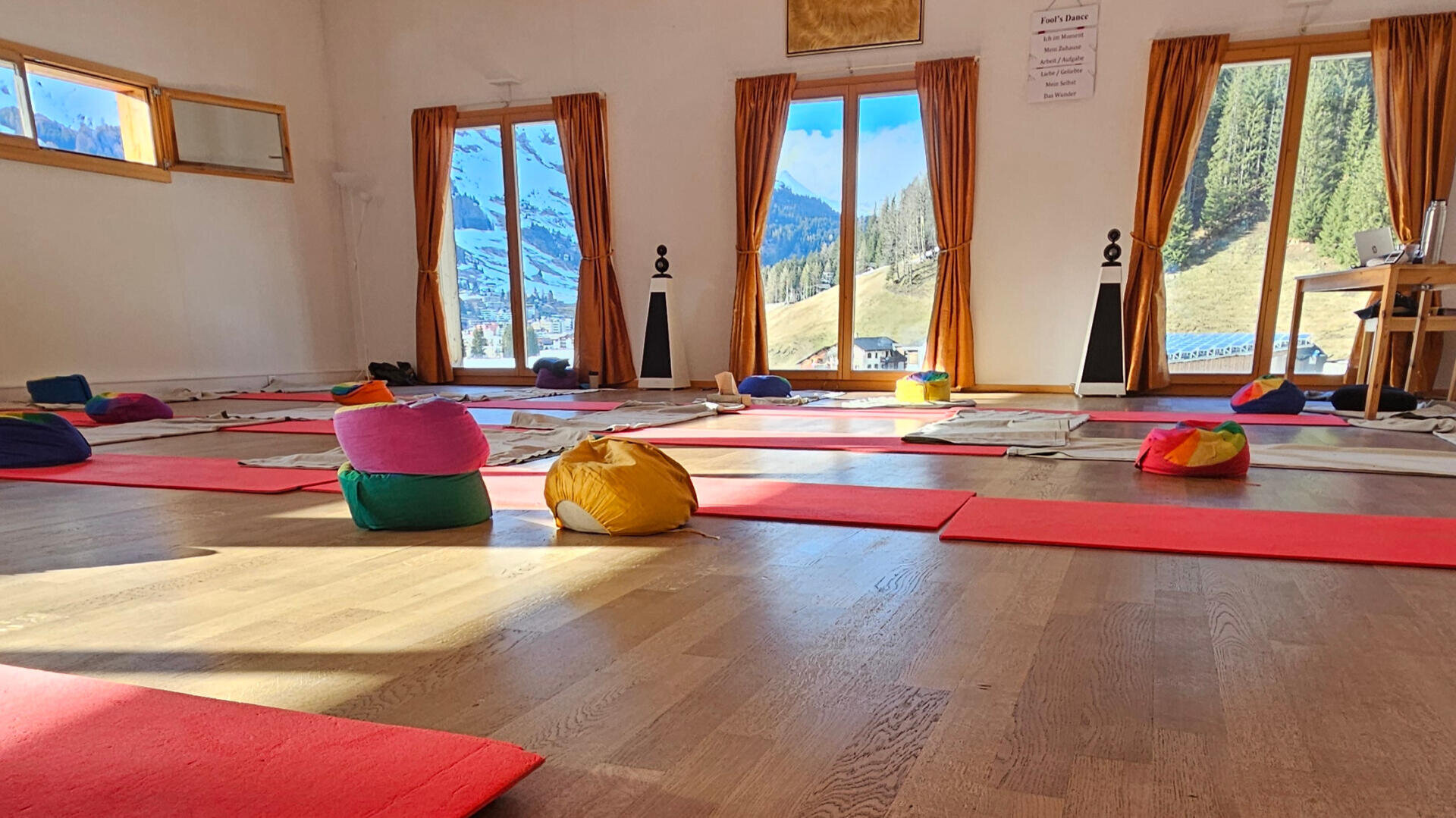 Ein sonnendurchflutetes Yogastudio mit Holzböden, roten Matten und bunten Kissen. Große Fenster und Türen geben den Blick auf verschneite Berge und Kiefern frei und schaffen eine ruhige und einladende Atmosphäre.