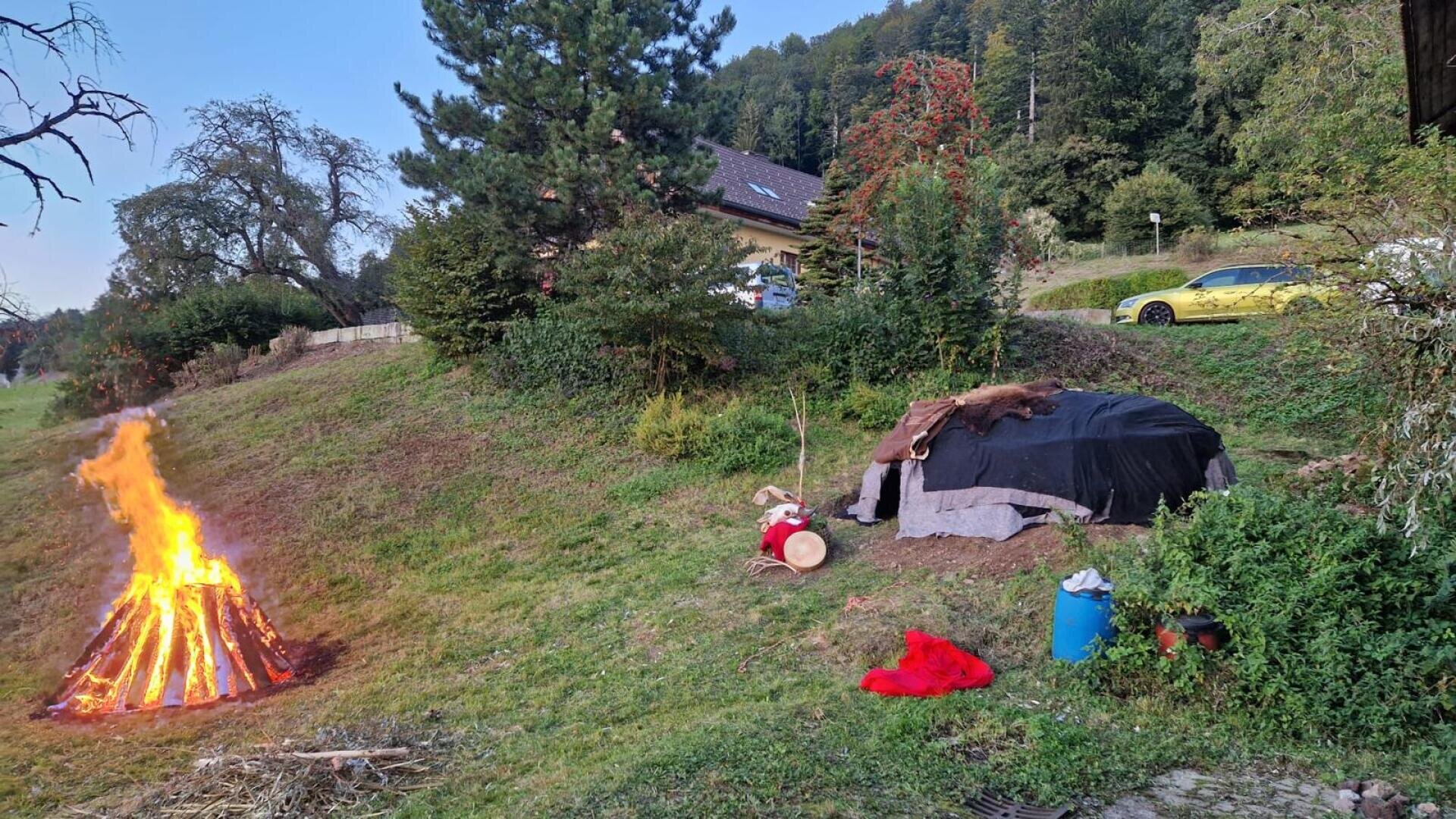 Ein Lagerfeuer brennt auf einem grasbewachsenen Hügel in der Nähe einer kleinen Hütte, die mit Decken und Tierfellen bedeckt ist. In der Nähe befinden sich Trommeln, ein rotes Tuch, ein blauer Eimer und Bäume; im Hintergrund parkt ein gelbes Auto neben einem Haus.