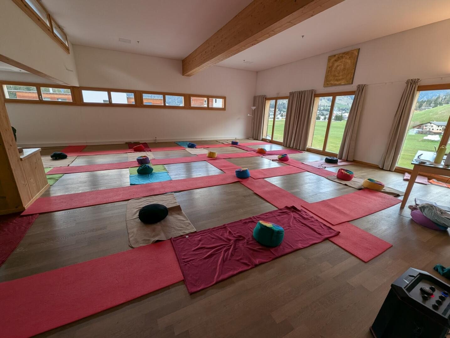 Ein großer, heller Raum, eingerichtet für eine Yogastunde mit roten Matten, Kissen und Handtüchern, die ordentlich auf dem Holzboden liegen. Die Fenster blicken auf eine Graslandschaft und Hügel und lassen natürliches Licht herein.