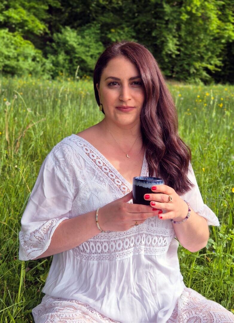 Eine Frau mit langen braunen Haaren sitzt in einem grasbewachsenen Feld, trägt ein weißes Spitzenkleid und hält eine dunkle Tasse. Sie trägt roten Nagellack und ist von grünem Laub und gelben Wildblumen umgeben.