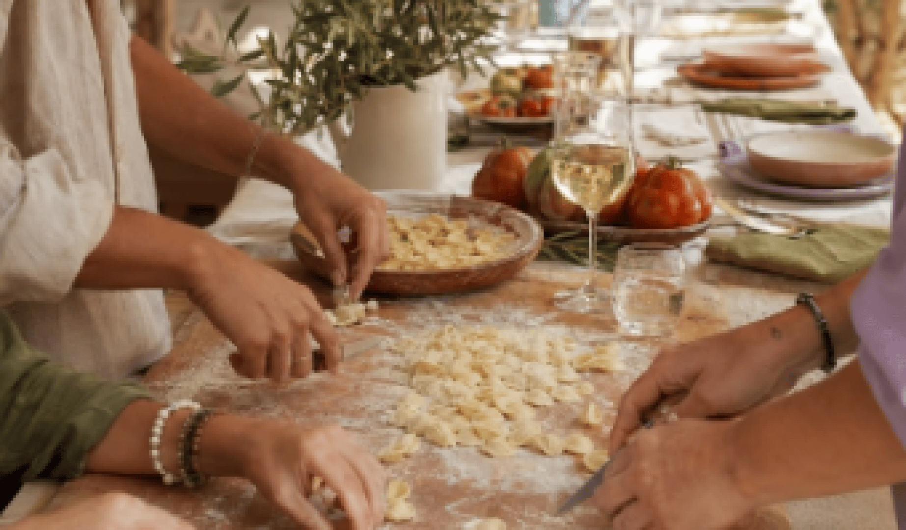 Drei Personen formen auf einer bemehlten Holzunterlage frischen Nudelteig. Im Hintergrund sind Weingläser, Tomaten und ein gedeckter Esstisch zu sehen, was auf die gemeinsame Zubereitung einer Mahlzeit hindeutet.