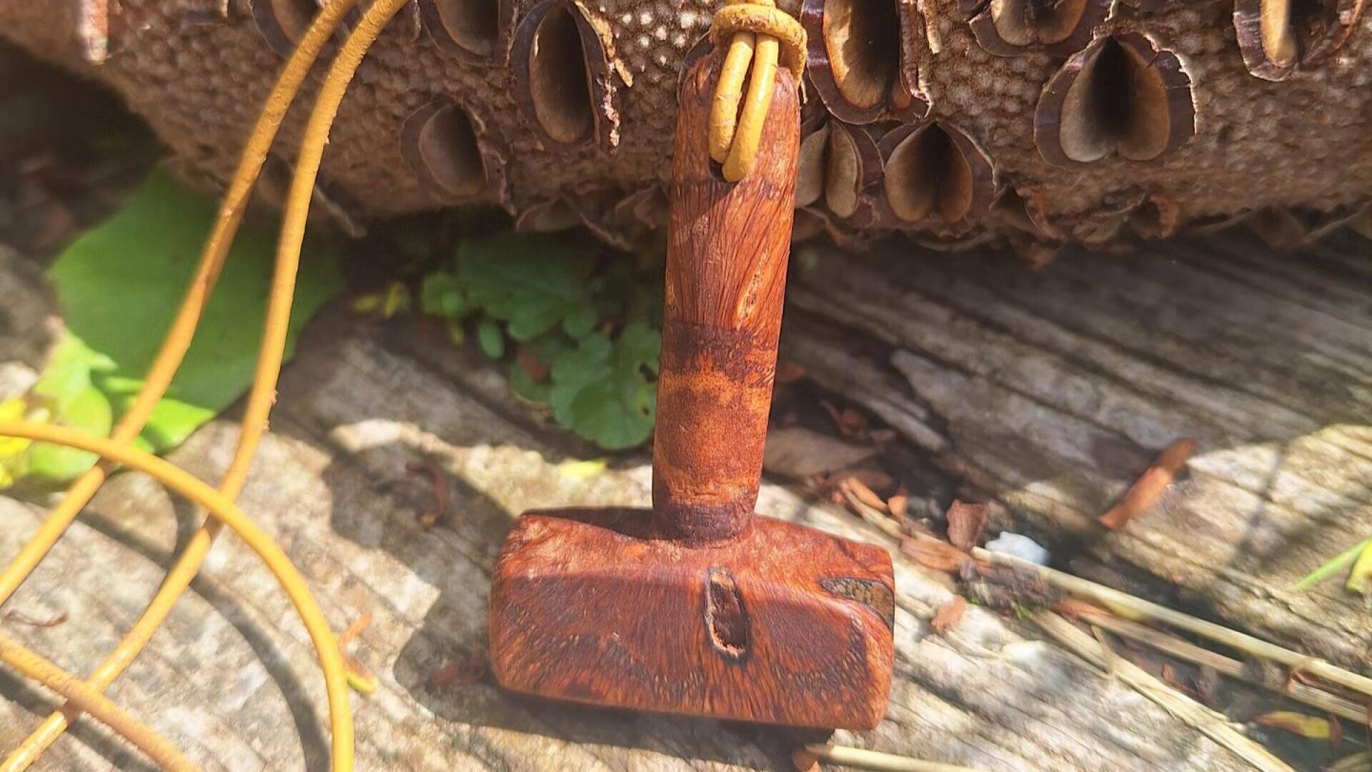 An einer Lederkordel hängt ein hölzerner Anhänger in Form eines Hammers, der auf einer verwitterten Holzfläche platziert ist, im Hintergrund sind braune Samenkapseln und grüne Blätter zu sehen.