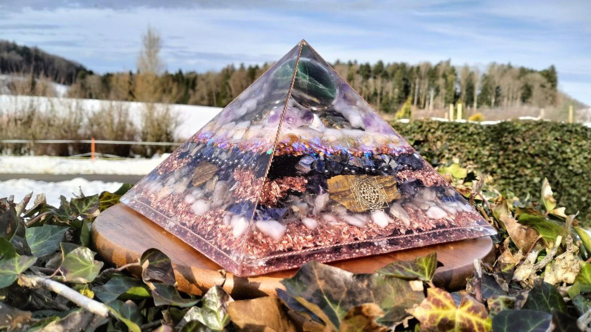 Eine farbenfrohe Orgonitpyramide mit eingebetteten Steinen und Metallen steht auf einem Holzsockel im Freien, umgeben von grünem Efeu und einer winterlichen Landschaft mit Wald im Hintergrund.