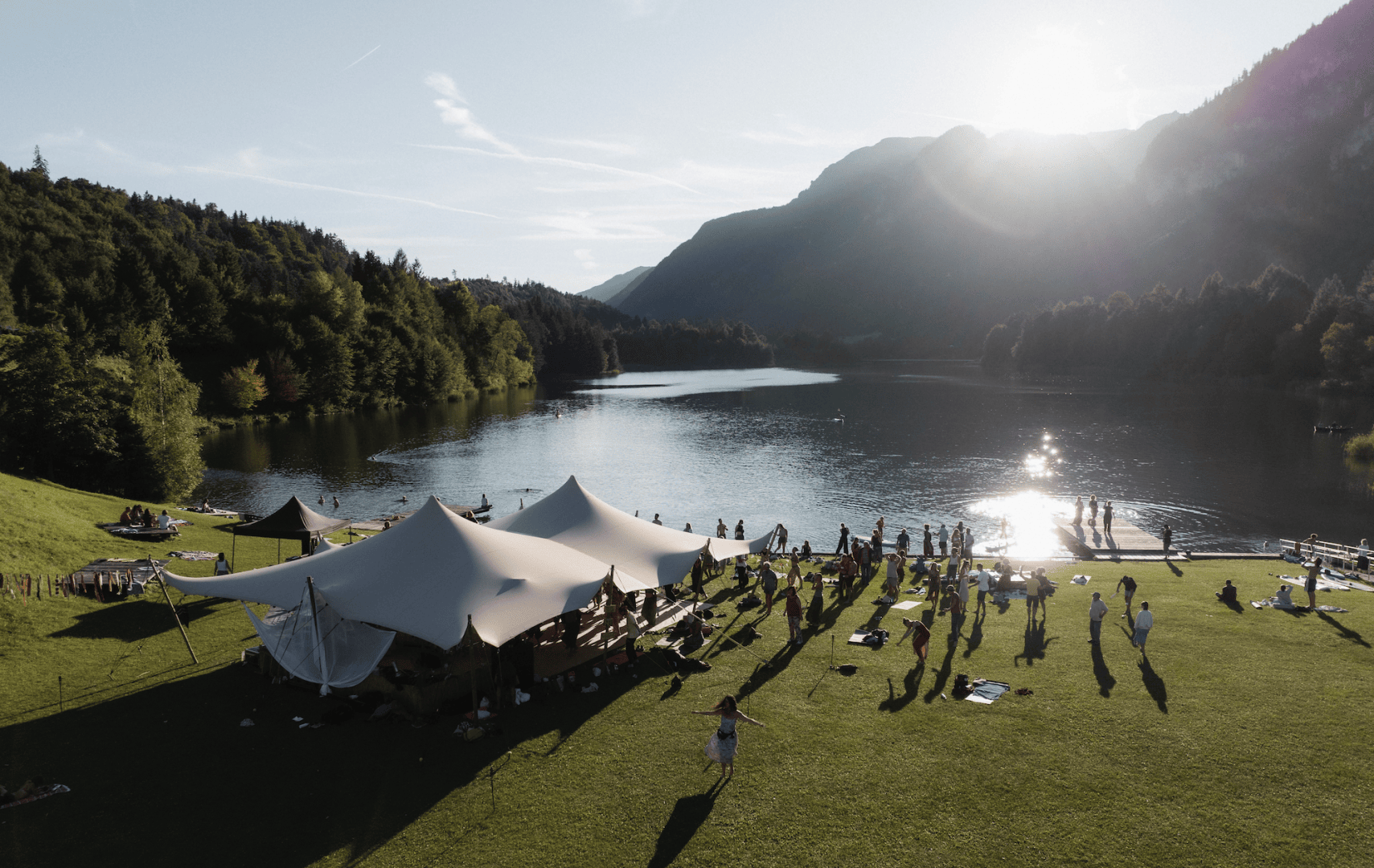 Menschen versammeln sich an einem grasbewachsenen Seeufer in der Nähe eines großen Zeltes mit Bergen und Bäumen im Hintergrund. Die Sonne scheint von oben, wirft lange Schatten und schafft eine helle, heitere Atmosphäre am Wasser.