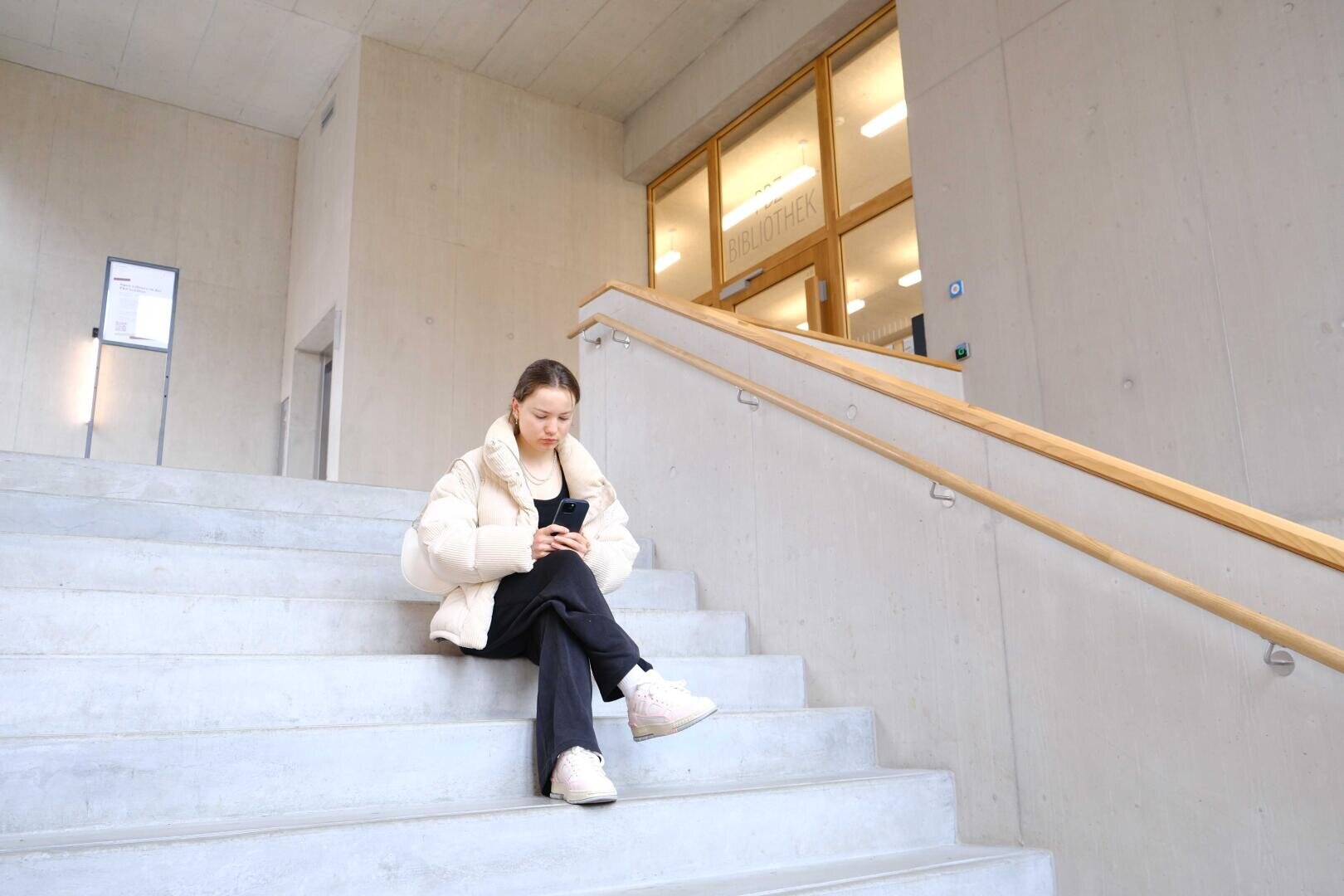 Eine Person in einem weißen Kittel sitzt auf einer Betonstufe und schaut auf ihr Handy. Im Hintergrund ist ein modernes Gebäude mit großen Fenstern und einem Schild mit der Aufschrift „BIBLIOTHEK“ zu sehen.
