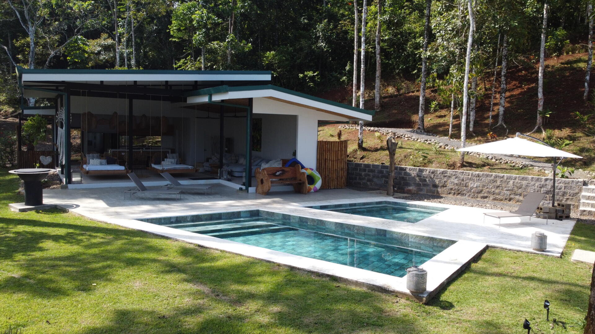 Eine moderne Villa am Pool mit offenem Wohnbereich, Loungesesseln und einer überdachten Terrasse, eingebettet in eine üppige, grüne Tropenlandschaft mit hohen Bäumen und einer Steinmauer im Hintergrund.