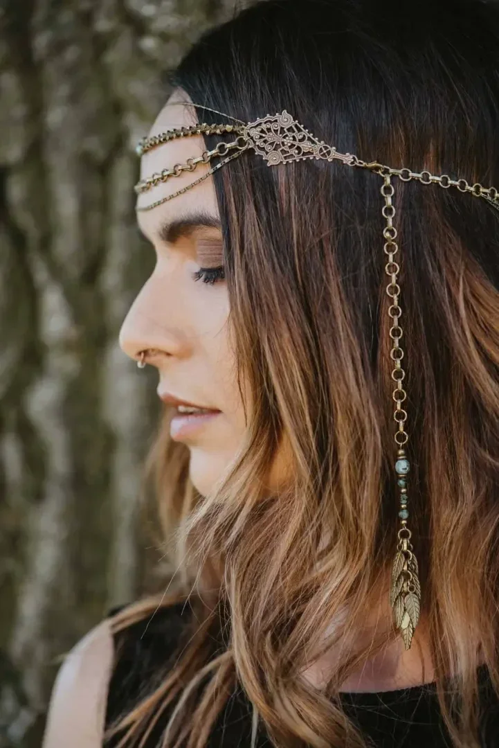 Eine Person mit welligem braunem Haar trägt einen verzierten goldenen Kopfschmuck aus einer Kette mit einem Blattanhänger und Perlen und steht vor einem Baum.
