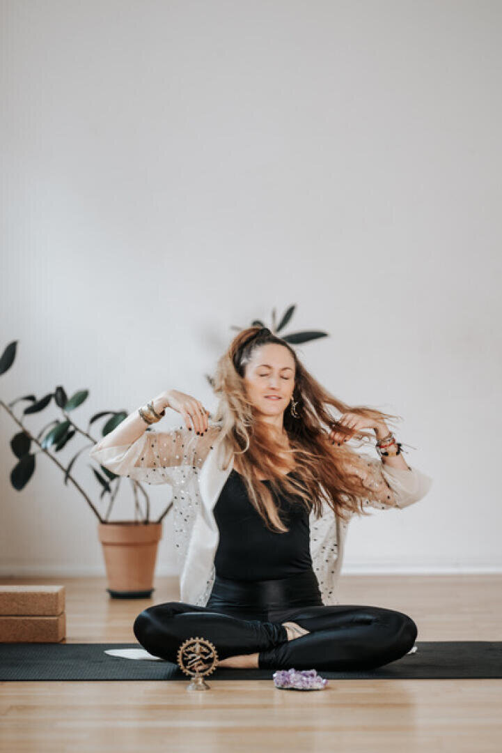 Eine Frau mit langen Haaren sitzt im Schneidersitz auf einer Yogamatte in einem Haus, die Augen geschlossen und die Hände leicht erhoben. Sie trägt ein schwarzes Outfit mit einer hellen Strickjacke. Im Hintergrund stehen eine Topfpflanze und Yogablöcke.
