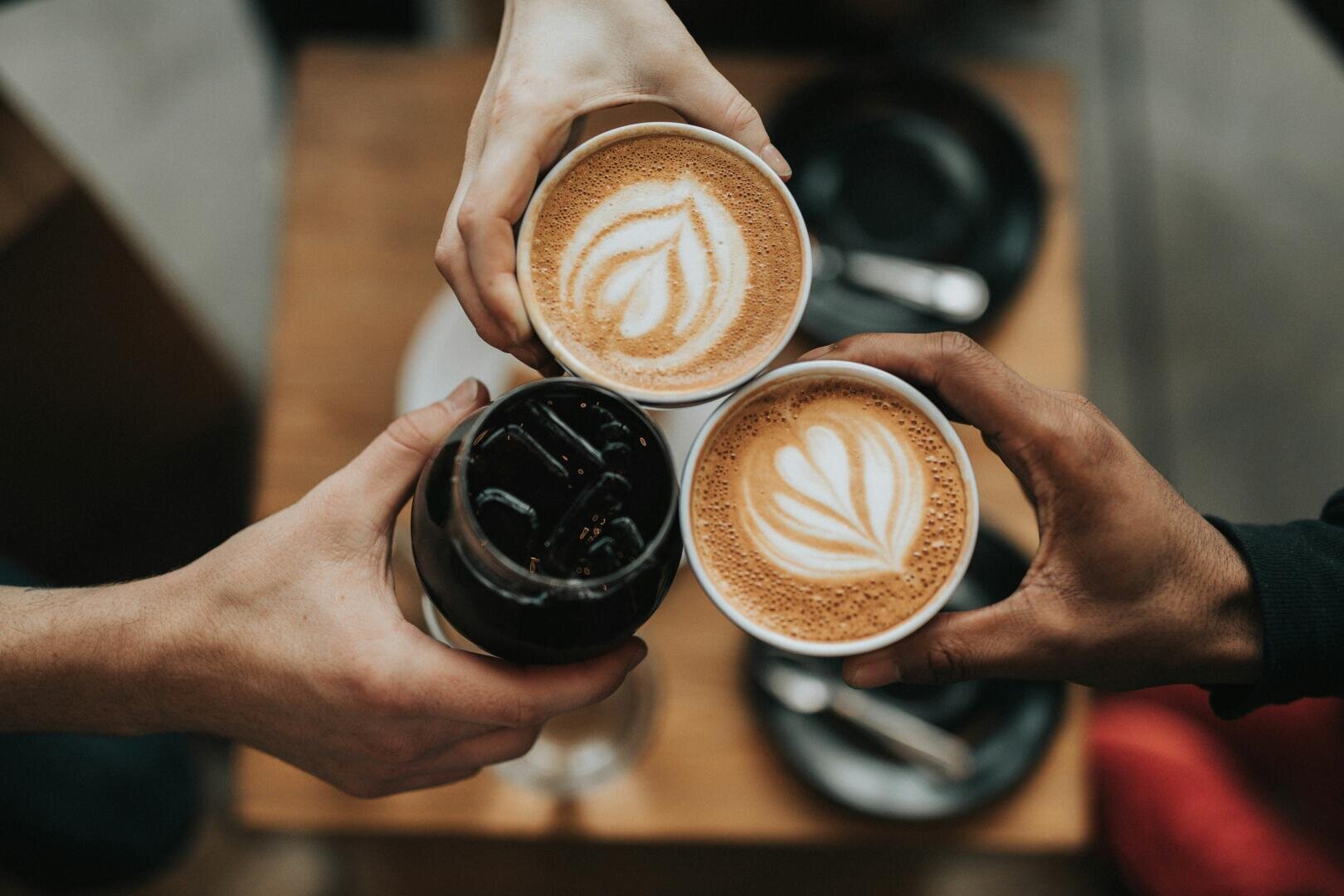 Drei Hände halten zwei Lattes mit herzförmigem Schaummuster und ein Glas Eiskaffee in einer Anstoßgeste.