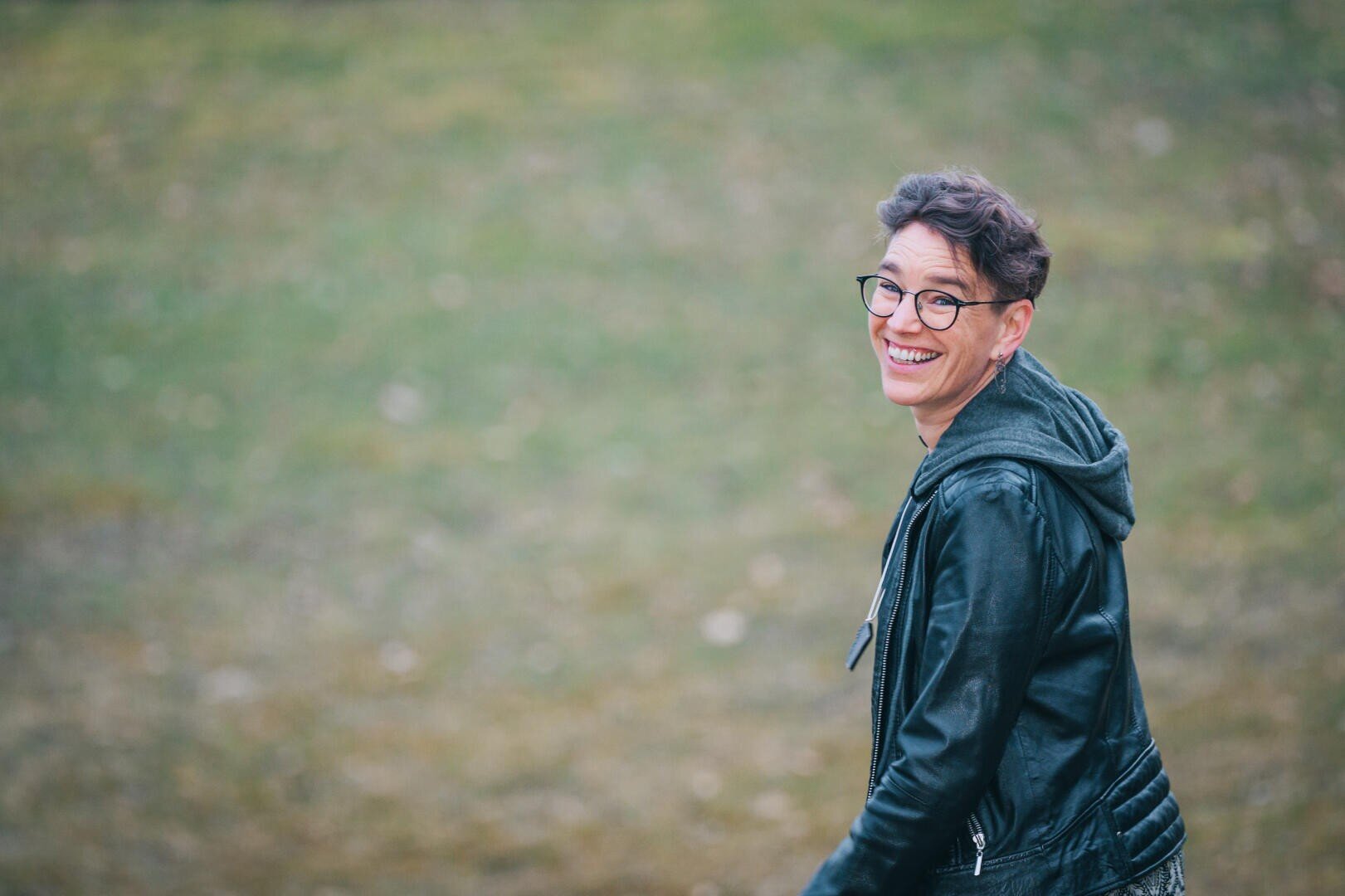 Eine Person, die eine Brille und eine schwarze Lederjacke trägt, lächelt, während sie über die Schulter zurückblickt; sie steht draußen auf einer Rasenfläche.