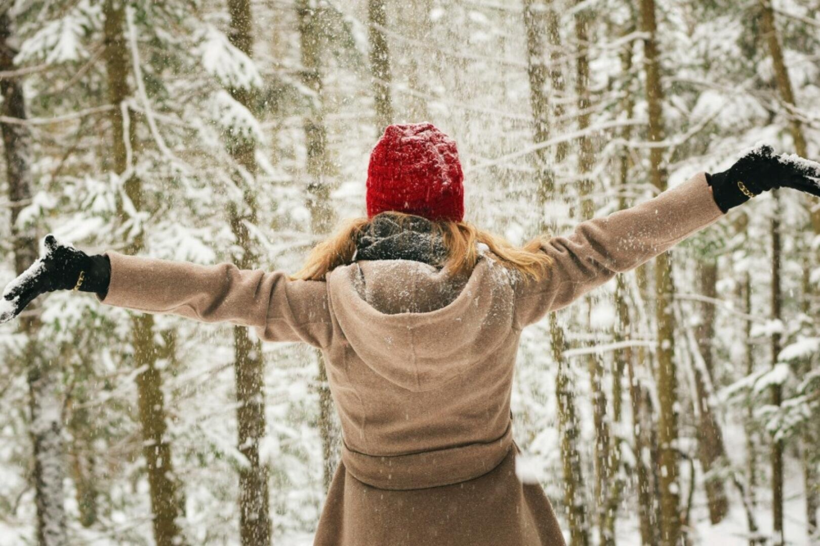 Eine Person mit rotem Hut und hellbraunem Mantel steht mit ausgestreckten Armen in einem verschneiten Wald, während um sie herum sanft Schnee fällt.