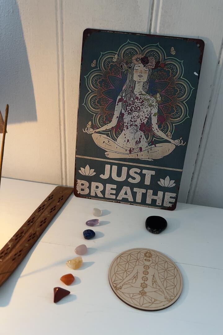 Auf einer weißen Fläche steht eine Holztafel mit der Abbildung einer meditierenden Frau und der Aufschrift „JUST BREATHE“. Daneben liegen Räucherstäbchen, ein Untersetzer mit kreisförmigem Muster, ein schwarzer Stein und sechs bunte Kristalle, die in einer Reihe angeordnet sind.