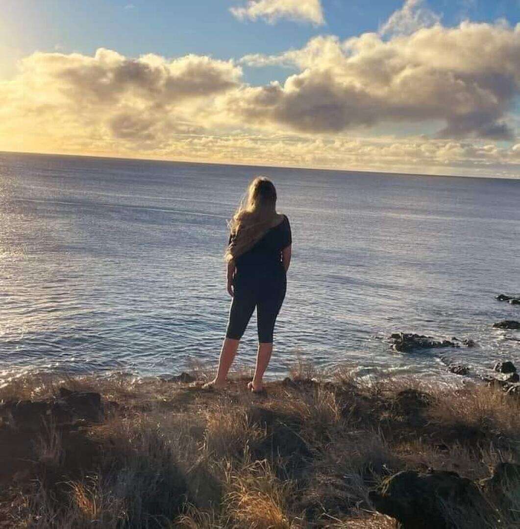 Eine Person mit langen Haaren steht auf einer grasbewachsenen Klippe mit Blick auf das Meer bei Sonnenuntergang, wobei sich das goldene Sonnenlicht auf dem Wasser und den Wolken am Himmel spiegelt.