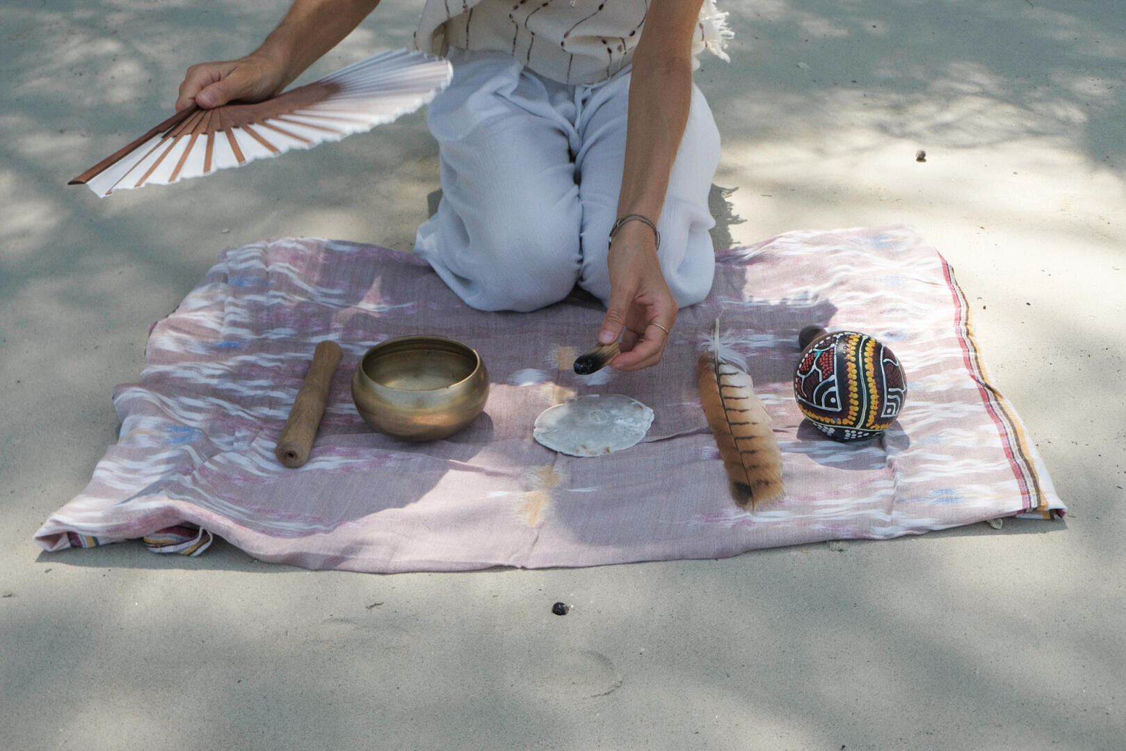 Eine Person kniet im Freien auf einem Tuch und hält einen weißen Fächer und einen kleinen Gegenstand in der Hand. Vor ihr liegen eine Messingschale, ein Holzstab, eine Feder, eine Muschel und ein bunter Stoffball, die für ein Ritual oder eine Zeremonie arrangiert wurden.