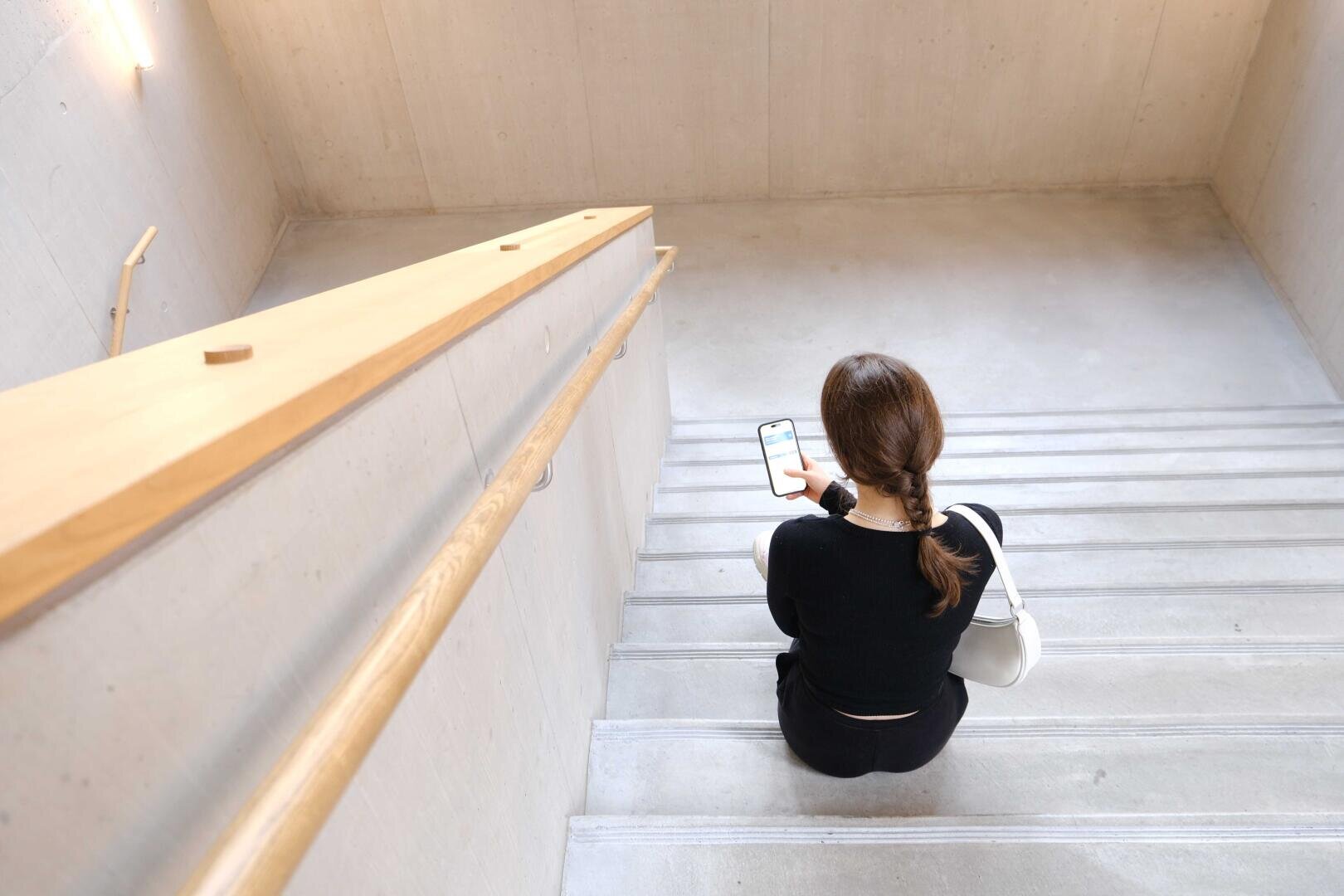 Eine Person mit einem Zopf, ganz in Schwarz gekleidet, sitzt auf einer Betontreppe in einem modernen Gebäude und schaut auf ein Smartphone.