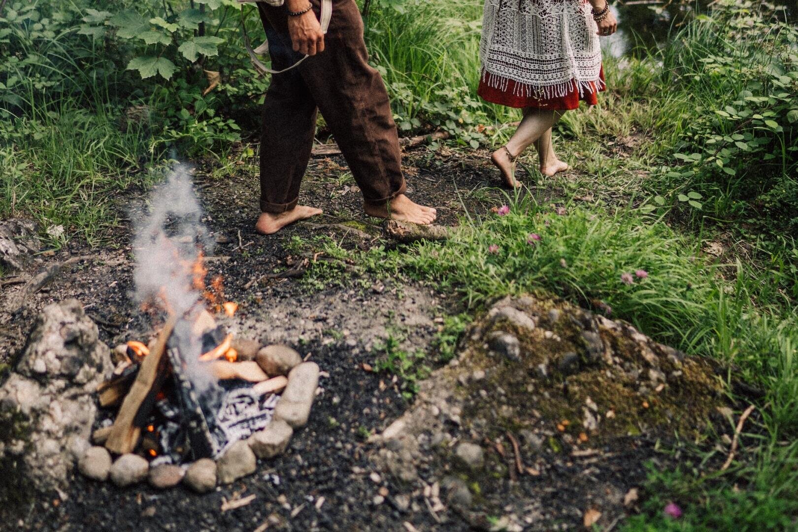 Zwei barfüßige Menschen gehen in einer grasbewachsenen, bewaldeten Gegend um ein kleines, von Steinen umgebenes Lagerfeuer herum. Eine Person trägt eine braune Hose, die andere ein gemustertes Kleid, und die Szene ist von Grün umgeben.