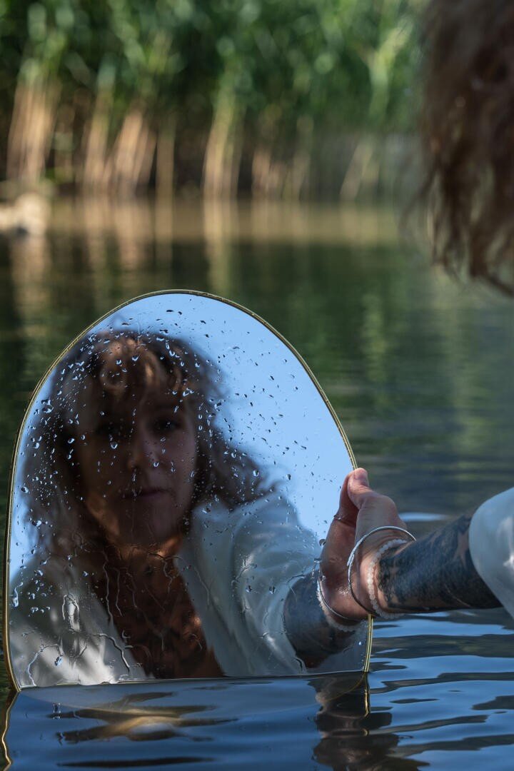 Eine Person hält einen Spiegel über das Wasser, sodass ihr Spiegelbild zusammen mit Wassertropfen auf dem Glas zu sehen ist. Im Hintergrund ist verschwommenes Grün zu erkennen.