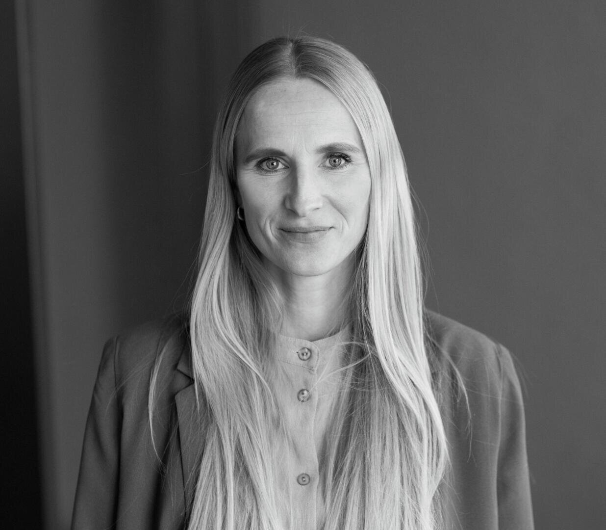 Schwarz-Weiß-Porträt einer Frau mit langen, glatten Haaren, die einen Blazer und ein zugeknöpftes Hemd trägt, vor einem schlichten Hintergrund steht und mit einem leichten Lächeln direkt in die Kamera blickt.