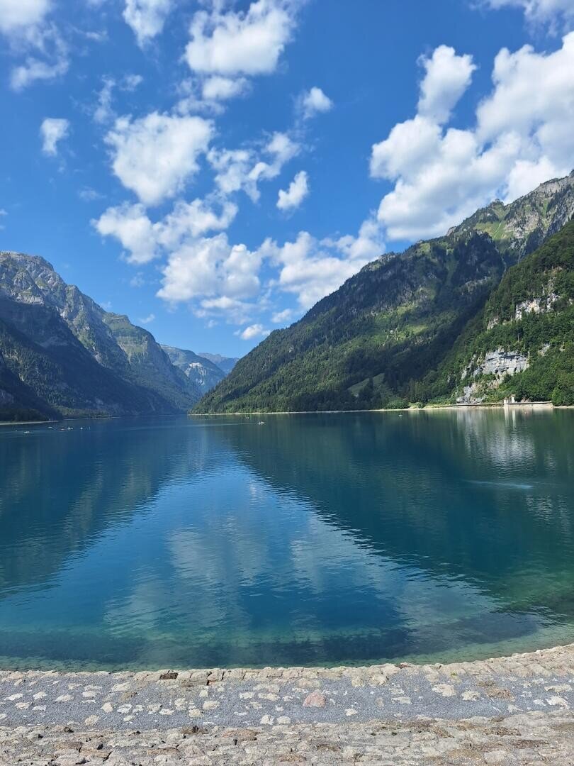 Ein kristallklarer, blauer See, umgeben von steilen, bewaldeten Bergen unter einem blauen Himmel mit vereinzelten Wolken.