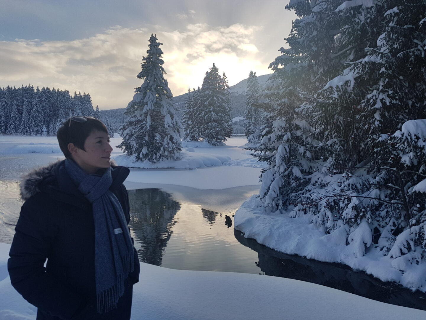 Eine Person in dunklem Wintermantel und Schal steht an einem verschneiten Flussufer, umgeben von schneebedeckten Bäumen und spiegelndem Wasser, während im Hintergrund die Sonne durch einen bewölkten Himmel scheint.