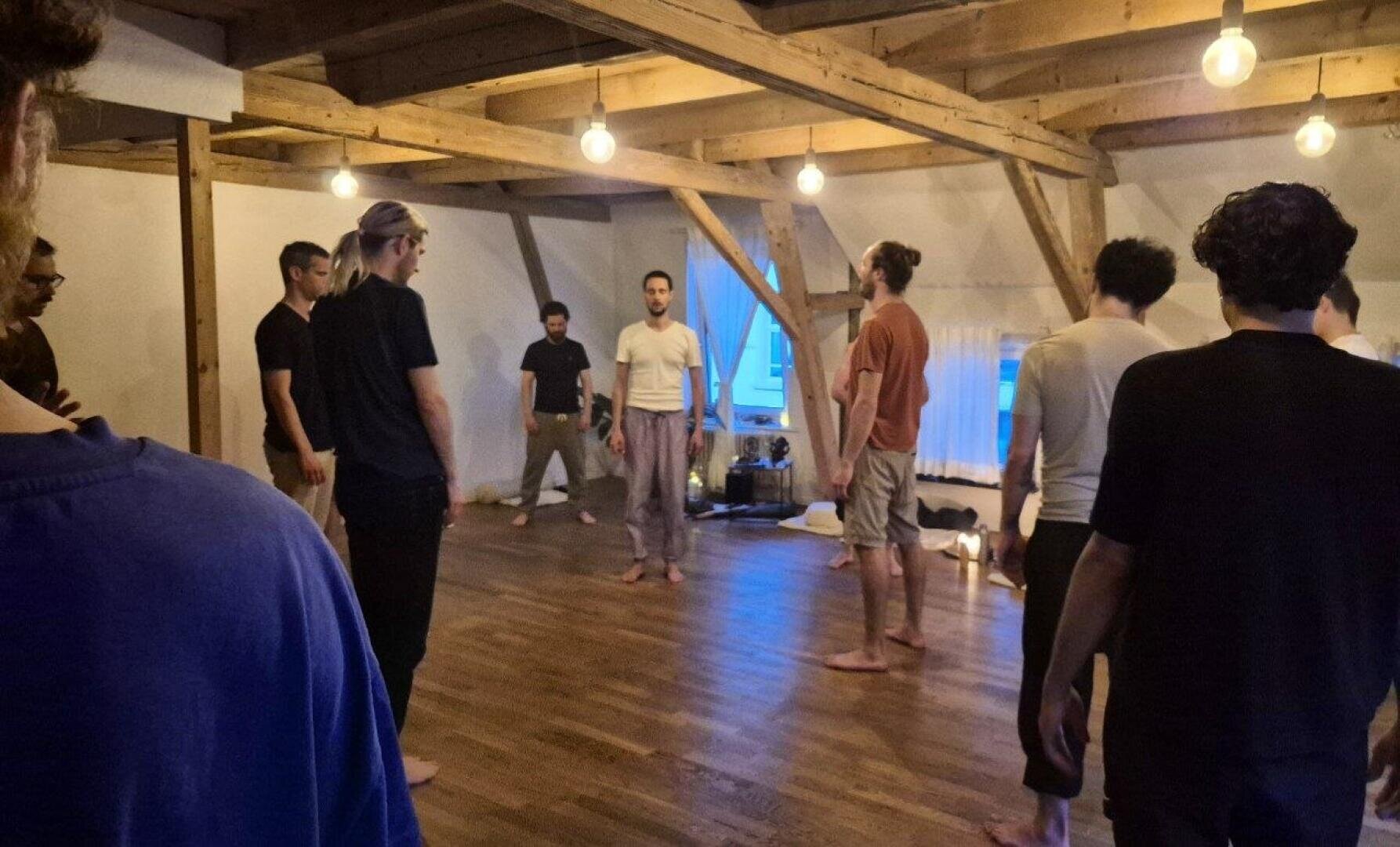 Eine Gruppe von Menschen steht barfuß im Kreis in einem hölzernen Raum mit freiliegenden Balken und hängenden Glühbirnen und nimmt an einer Indoor-Aktivität oder einem Workshop teil.