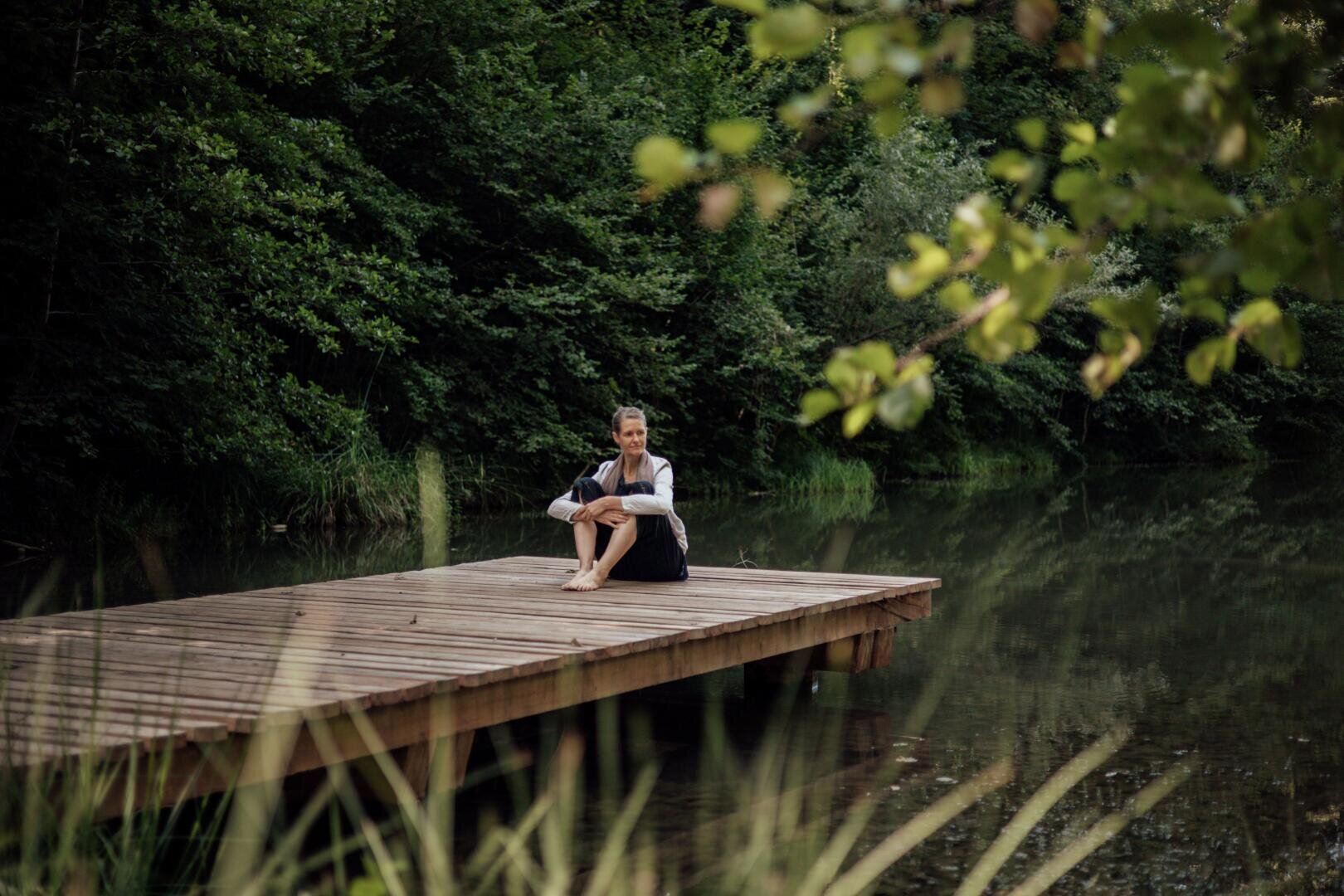 Eine Person sitzt im Schneidersitz auf einem hölzernen Steg über einem ruhigen See, umgeben von üppig grünen Bäumen und Pflanzen, vor dem Hintergrund einer friedlichen Naturlandschaft.