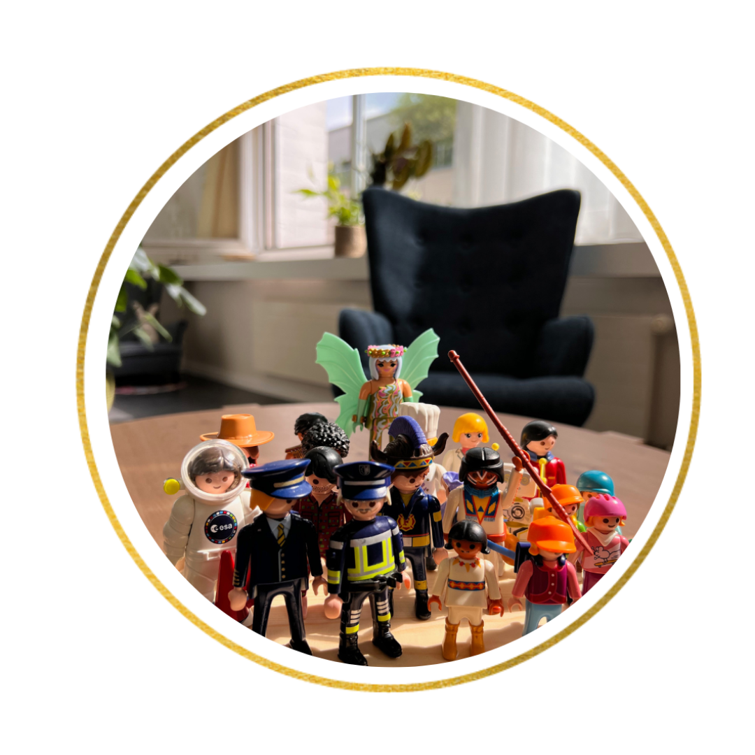 Eine Gruppe bunter Spielzeugfiguren, darunter eine Fee mit grünen Flügeln und verschiedene Figuren in Kostümen, ist auf einem Tisch in einem sonnendurchfluteten Raum angeordnet, im Hintergrund stehen ein Stuhl und Pflanzen.