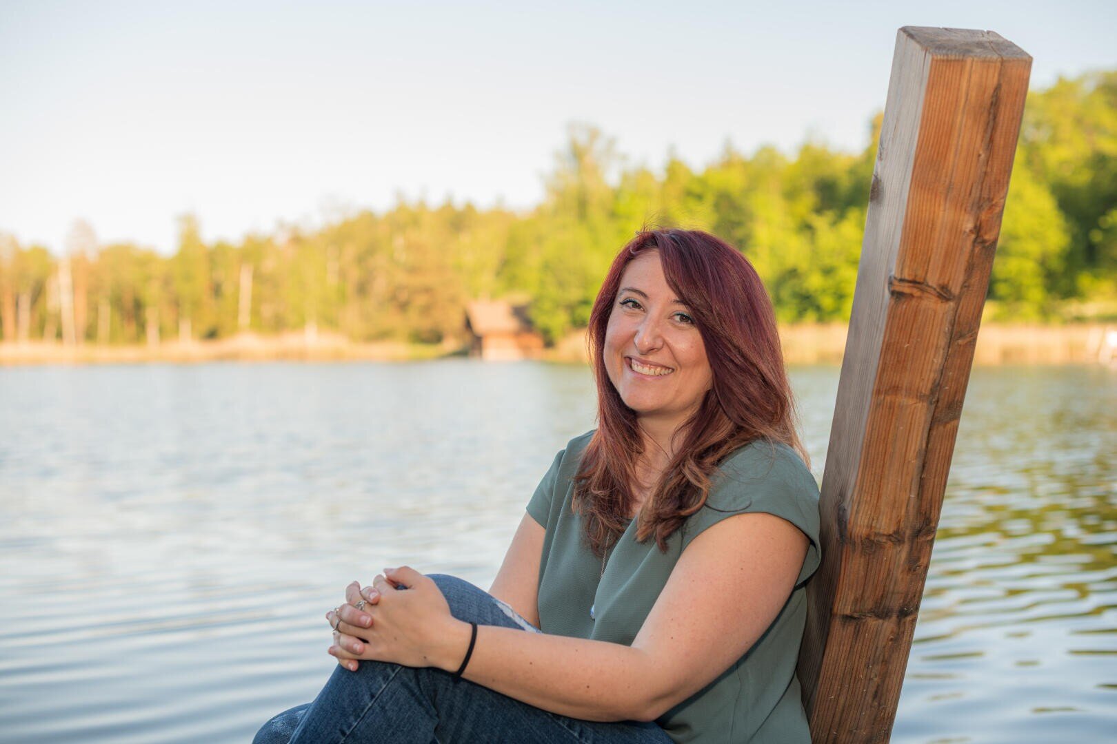 Eine Frau mit roten Haaren sitzt auf einem Holzsteg an einem See und lächelt in die Kamera. Im Hintergrund sind unter einem klaren Himmel Bäume und eine kleine Hütte zu sehen.