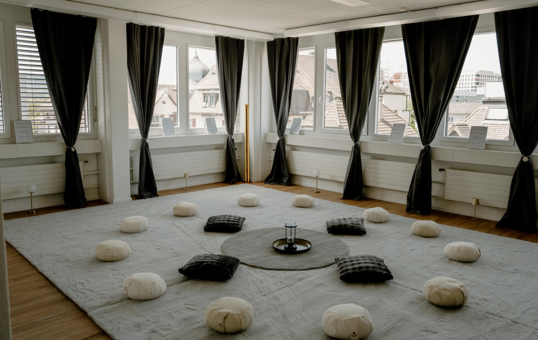Ein ruhiger Meditationsraum mit einem runden Teppich, einer Sanduhr in der Mitte und Bodenkissen, die kreisförmig auf einem großen Teppich angeordnet sind.