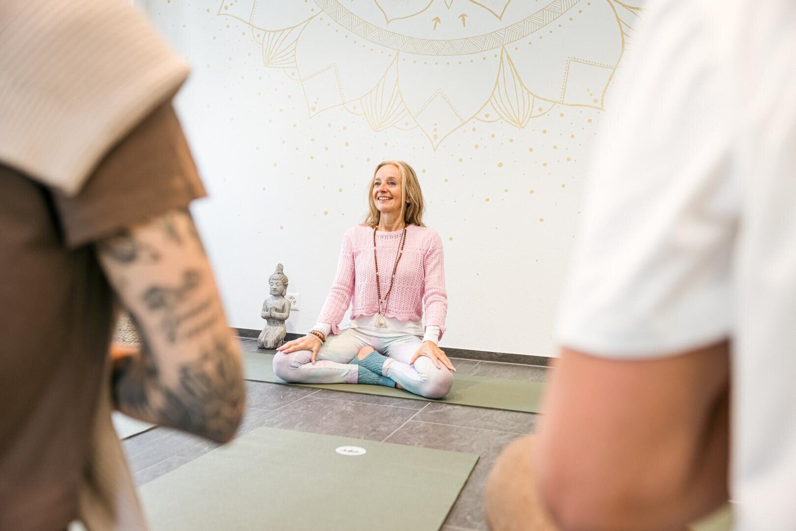 Eine Frau sitzt mit gekreuzten Beinen auf einer Yogamatte und lächelt in einem ruhigen Studio, an dessen Wand ein Mandala-Motiv zu sehen ist. Im Vordergrund sind zwei Personen zu sehen, die ihr zuhören. Daneben steht eine kleine Buddha-Statue.