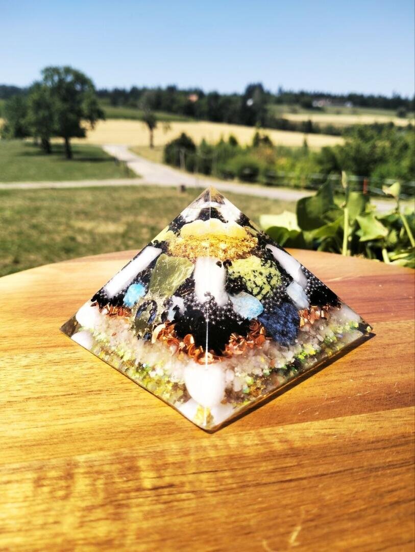 Eine bunte Pyramide aus Kunstharz mit Steinen und Metallspänen steht im Freien auf einer Holzunterlage, im Hintergrund sind grüne Felder, Bäume und ein blauer Himmel zu sehen.