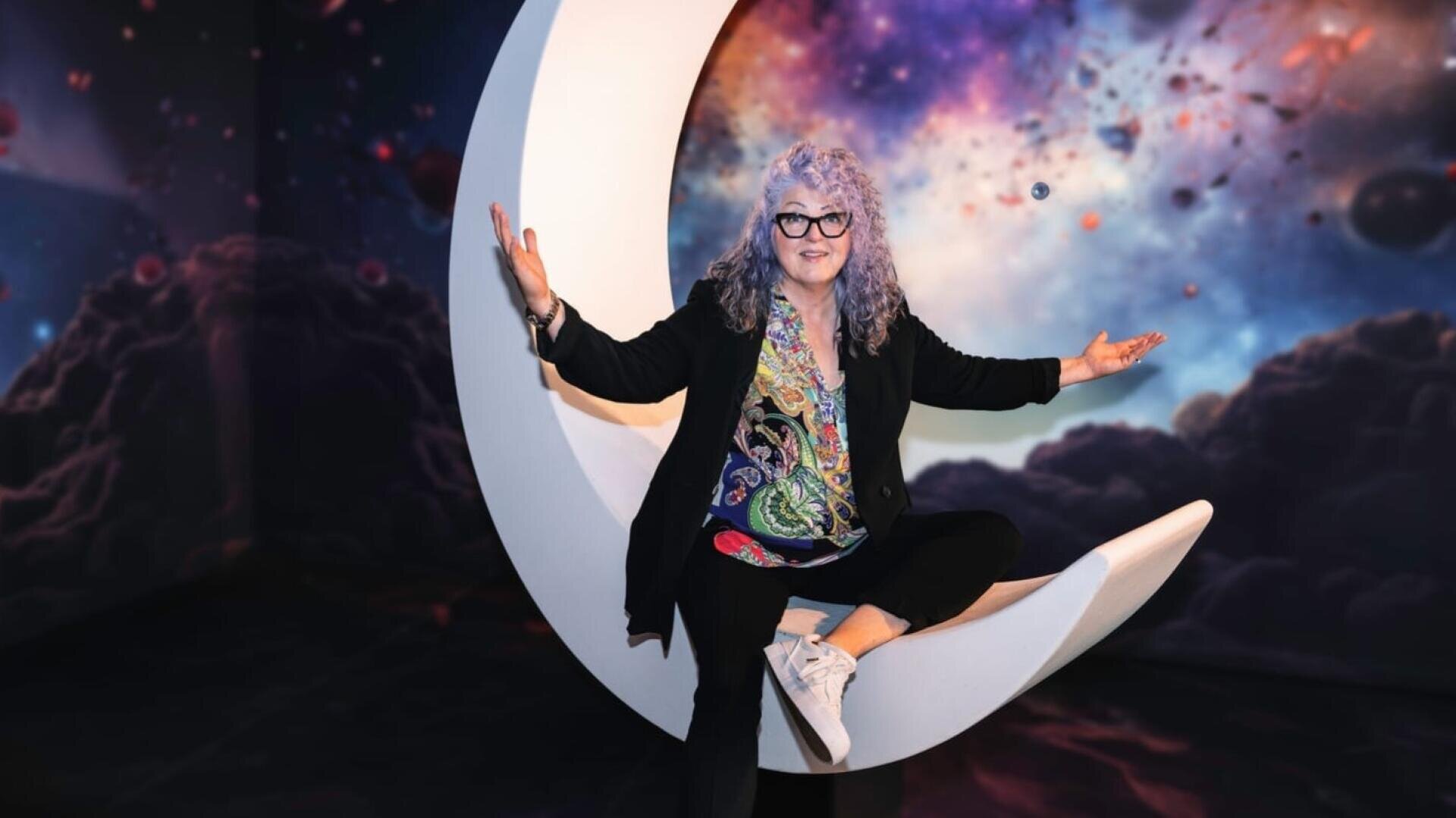 Eine Frau mit lockigem, grauem Haar, Brille und farbenfroher Kleidung sitzt auf einer großen Mondsichel-Skulptur, die Arme ausgestreckt, mit einem kosmischen, sternenübersäten Hintergrund hinter ihr.