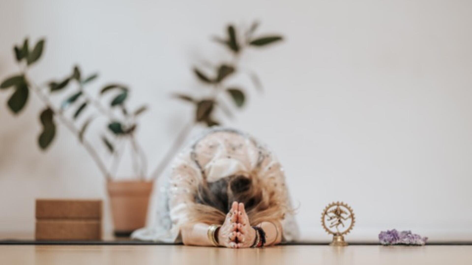 Eine Person in heller Kleidung nimmt auf einer Yogamatte die Haltung eines Kindes ein, die Hände zusammengedrückt, mit Zimmerpflanzen, Yogablöcken, einer kleinen Statue und lila Blumen im Hintergrund.
