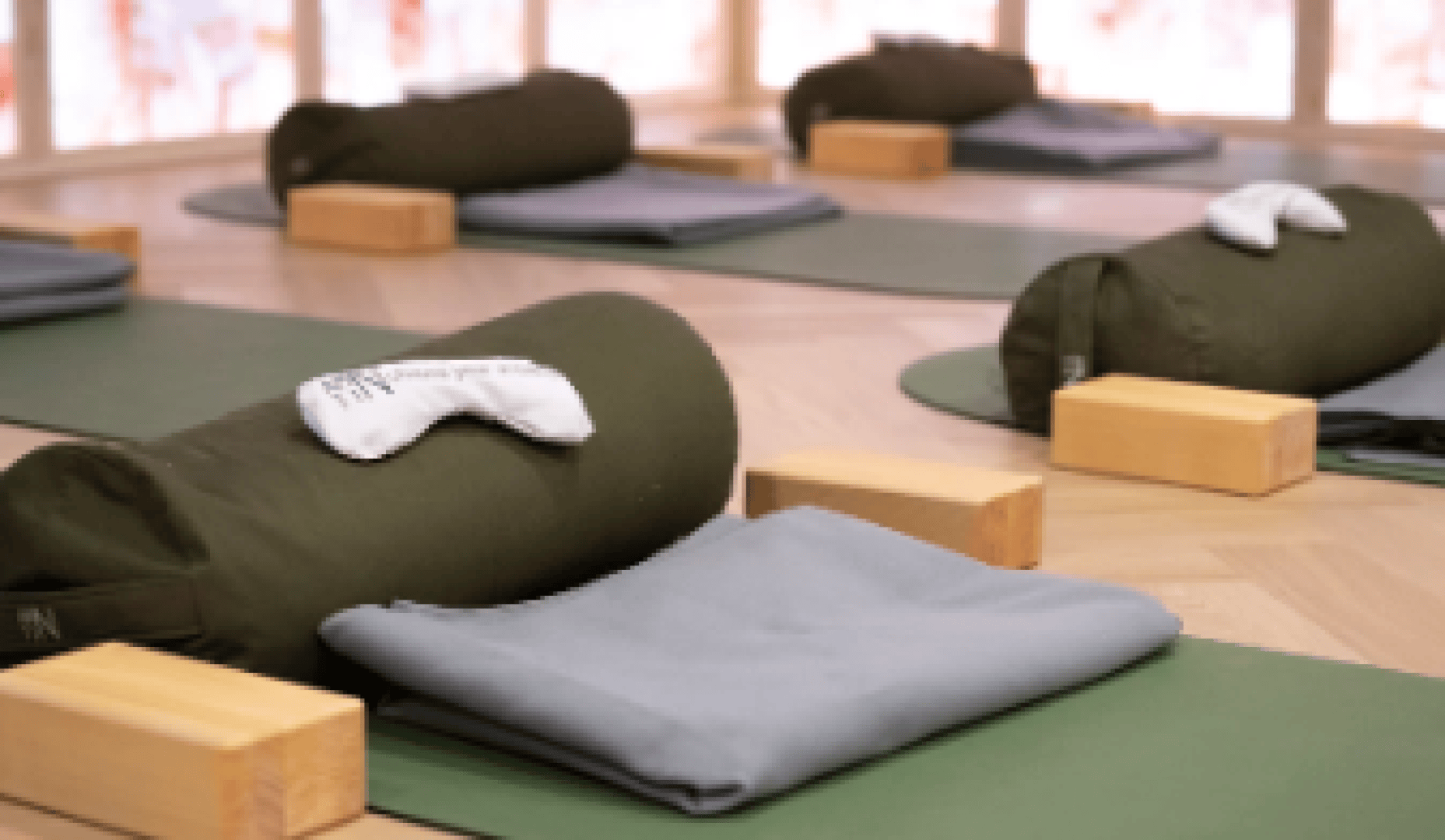 In einem hellen Studio sind Yogamatten mit Kissen, gefalteten Decken, Holzklötzen und Augenmasken ordentlich aufgereiht – bereit für einen Entspannungs- oder Meditationskurs.