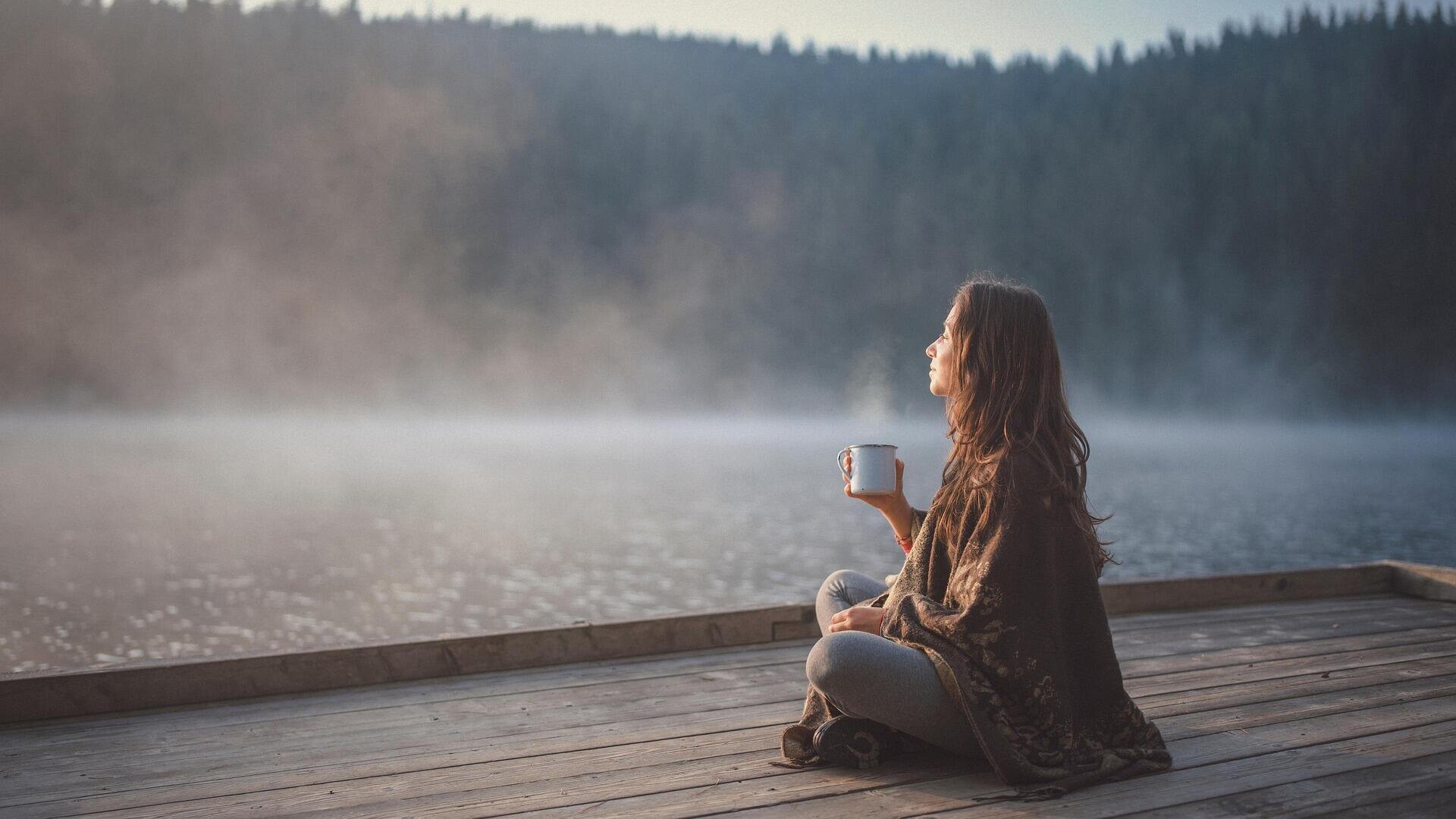 Eine Frau sitzt auf einem hölzernen Steg an einem nebligen See, hält einen Becher in der Hand und blickt friedlich auf das Wasser. Im Hintergrund sind kiefernbewachsene Hügel im sanften Licht des frühen Morgens zu sehen.