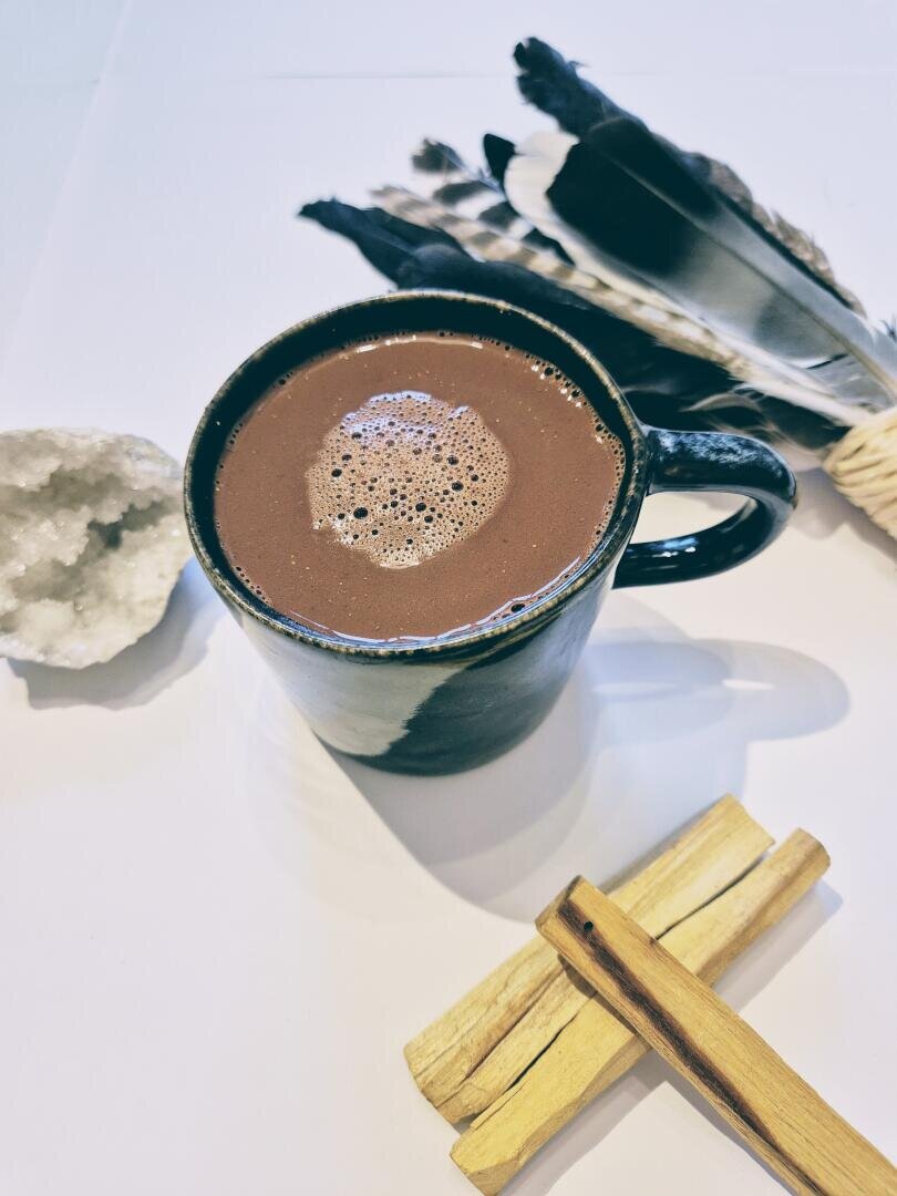 Ein schwarzer, mit heißer Schokolade gefüllter Becher steht auf einer weißen Fläche, daneben ein Federbündel, ein Geodenkristall und zwei Stücke Palo-Santo-Holz.