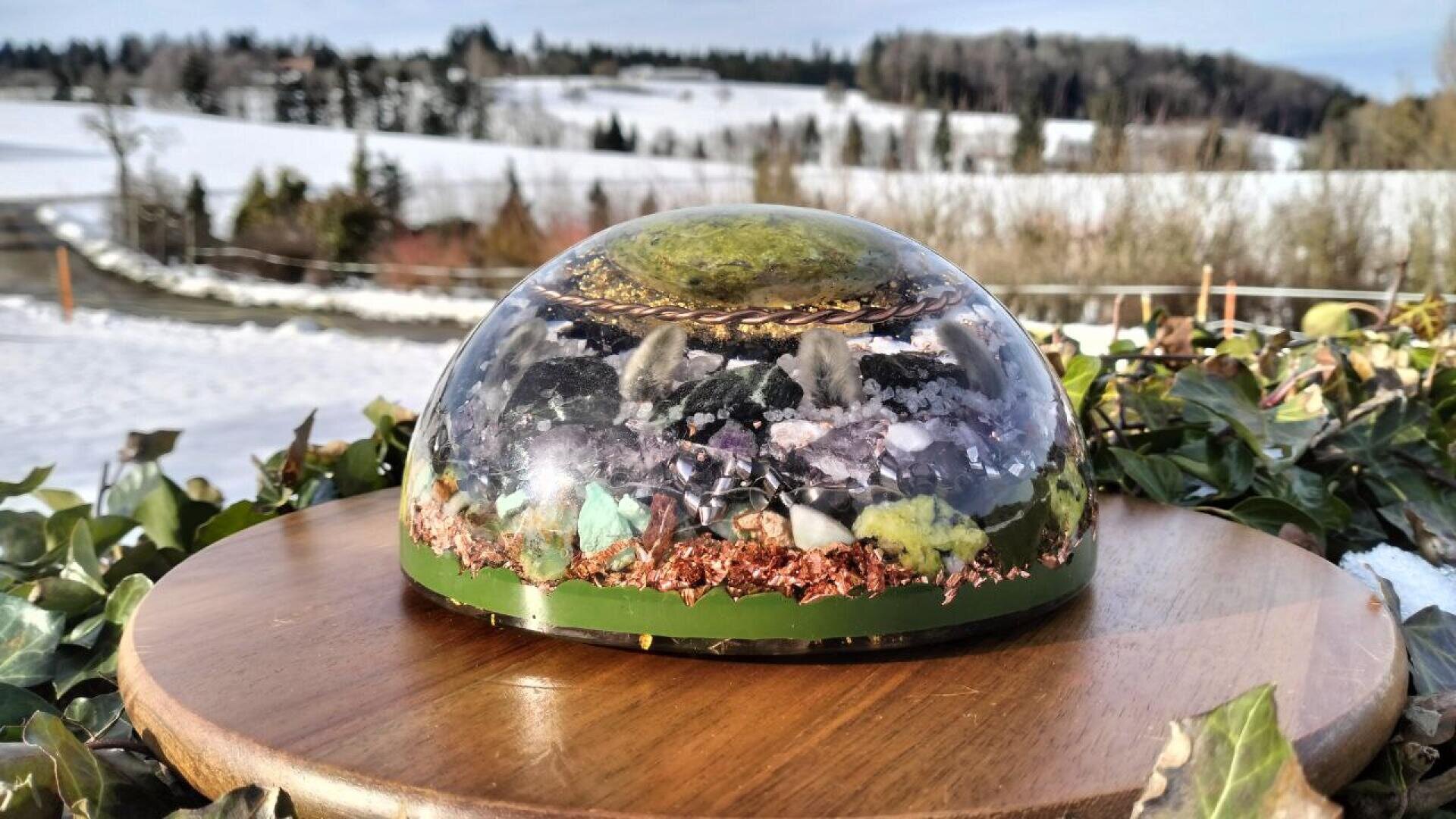 Eine kuppelförmige Orgonit-Skulptur, gefüllt mit verschiedenen Kristallen und Mineralien, steht auf einem Holzpodest im Freien, mit verschneiten Feldern, grünem Efeu und einer bewaldeten Landschaft im Hintergrund.