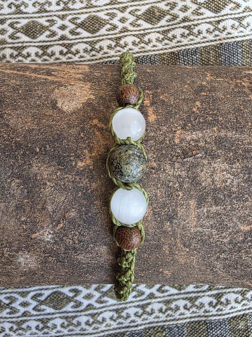 Ein handgefertigtes Armband mit grüner Flechtung, zwei weißen Perlen, einer grünen Perle mit grünen Sprenkeln in der Mitte und zwei kleinen braunen Perlen, präsentiert auf einer strukturierten braunen Oberfläche vor einem gemusterten Stoffhintergrund.