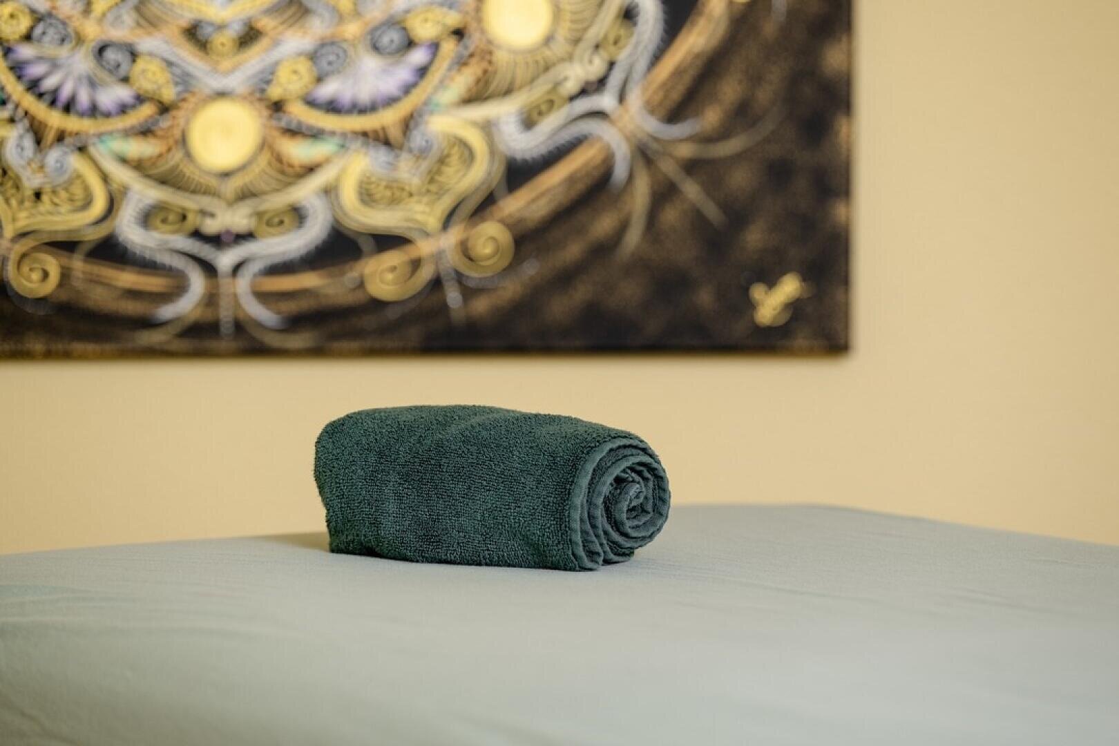 Auf einer hellen Massageliege liegt ein ordentlich aufgerolltes dunkelgrünes Handtuch, im Hintergrund hängt ein abstraktes Dekorationsgemälde an der Wand.