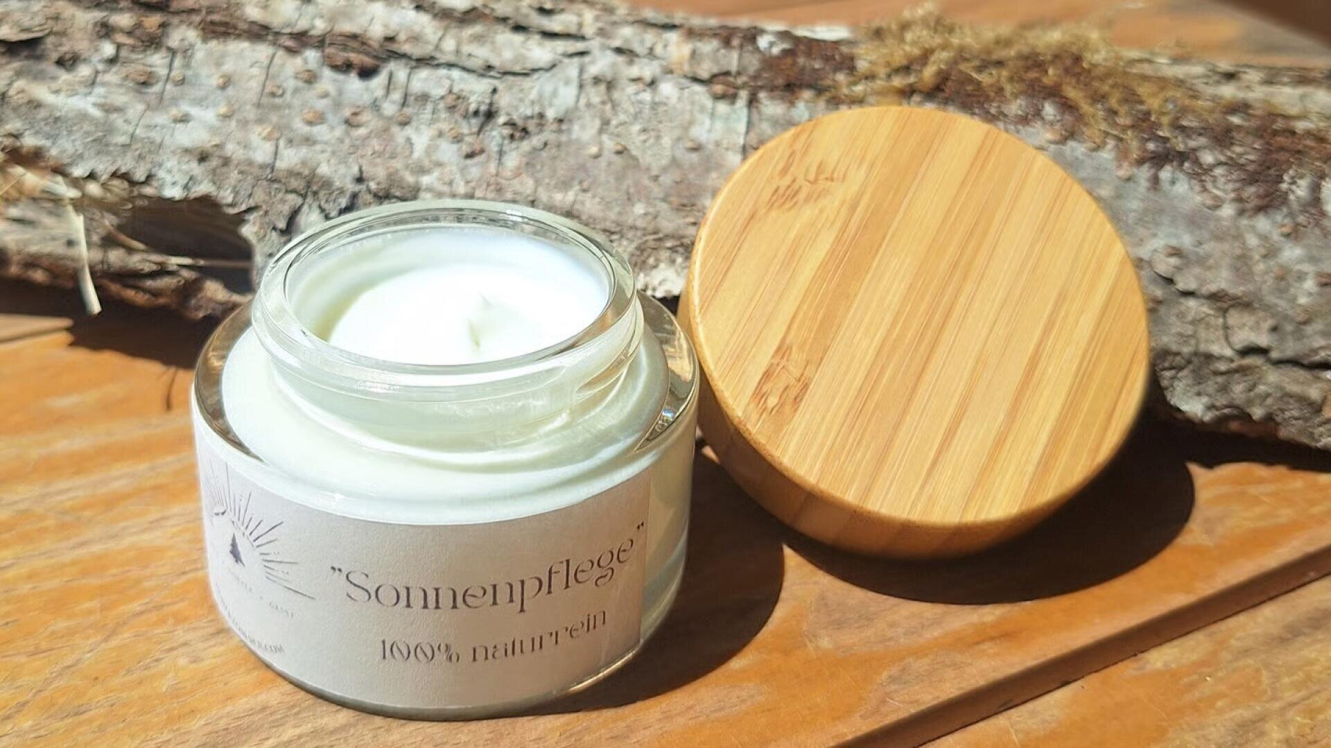 Ein offenes Glas mit weißer Creme, beschriftet mit „Sonnenpflege 100 % naturrein“, steht auf einer Holzunterlage neben einem Holzdeckel und einem Ast im Sonnenlicht.