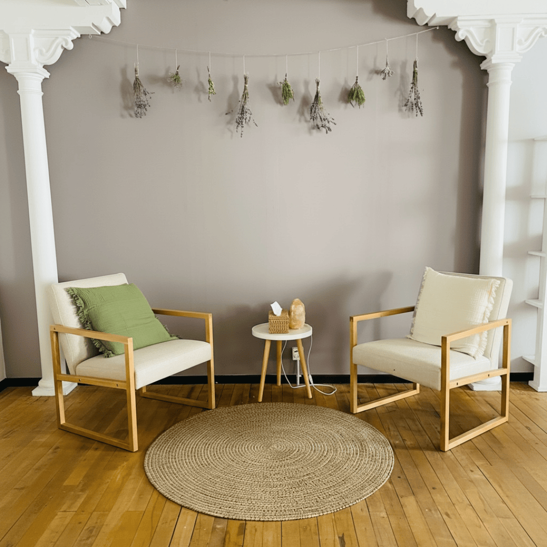 Zwei moderne Sessel mit Kissen stehen sich gegenüber, zwischen ihnen steht ein kleiner runder Tisch auf einem gewebten Teppich. An der Wand darüber hängen Trockenblumen, flankiert von weißen Ziersäulen. Die Szene wirkt gemütlich und einladend.
