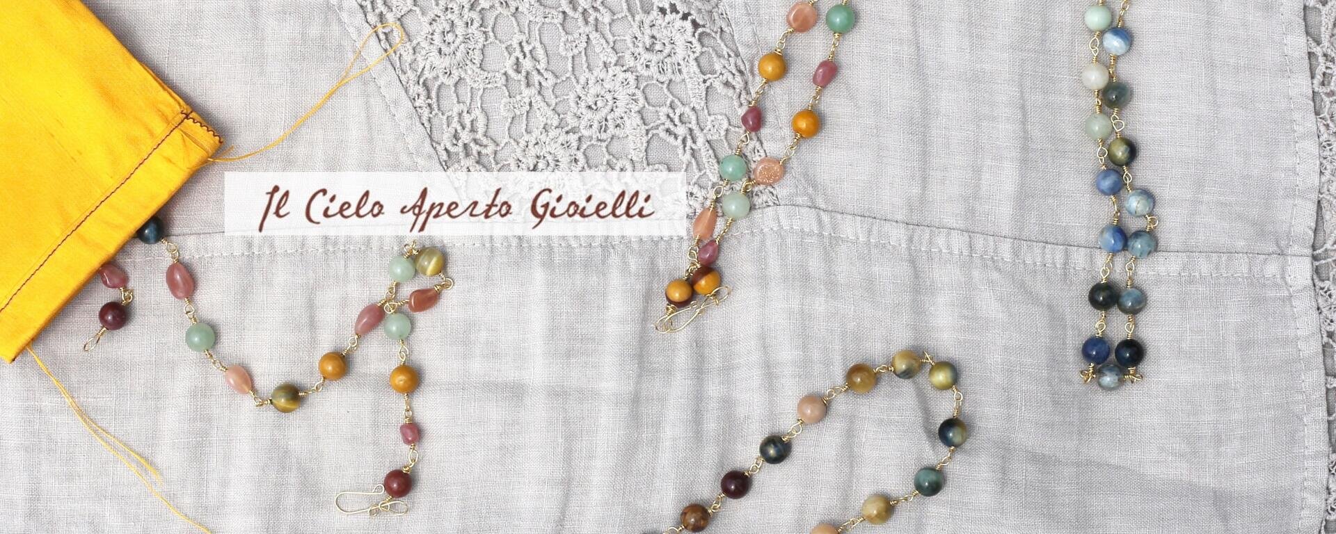 Farbenfrohe Perlenschmuckstücke sind auf einem hellen Stoff mit Spitzendetails präsentiert. Links befindet sich ein gelber Beutel. Der Schriftzug „Il Cielo aperto Gioielli“ ist auf einem weißen Streifen in der Mitte zu sehen.