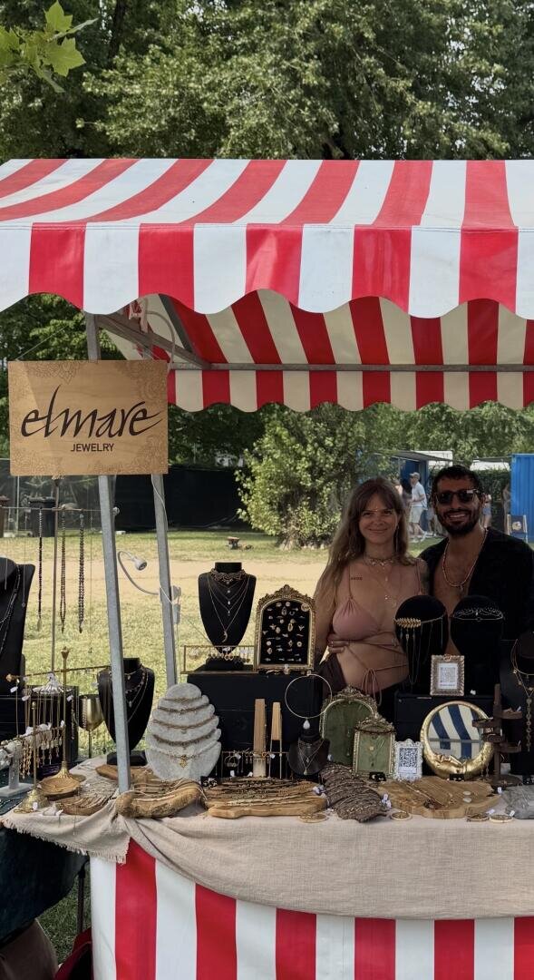 Hinter einem Schmuckstand unter einem rot-weiß gestreiften Zelt stehen zwei lächelnde Personen. Am Stand sind verschiedene Schmuckstücke ausgestellt sowie ein Holzschild mit der Aufschrift „elmave JEWELRY“. Im Hintergrund sind Bäume und Gras zu sehen.