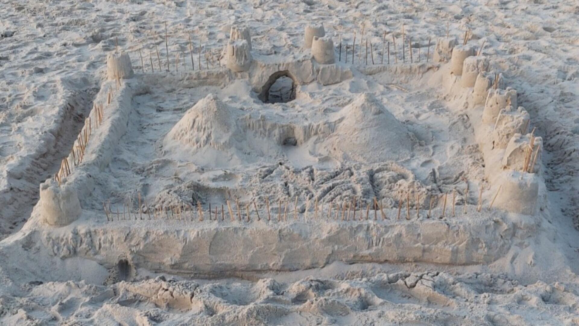 Eine detailreiche Sandburg mit Türmen, Mauern und Verzierungen aus Stöckchen, zwei zentralen Hügeln und einem gewölbten Eingang, gelegen an einem Sandstrand.
