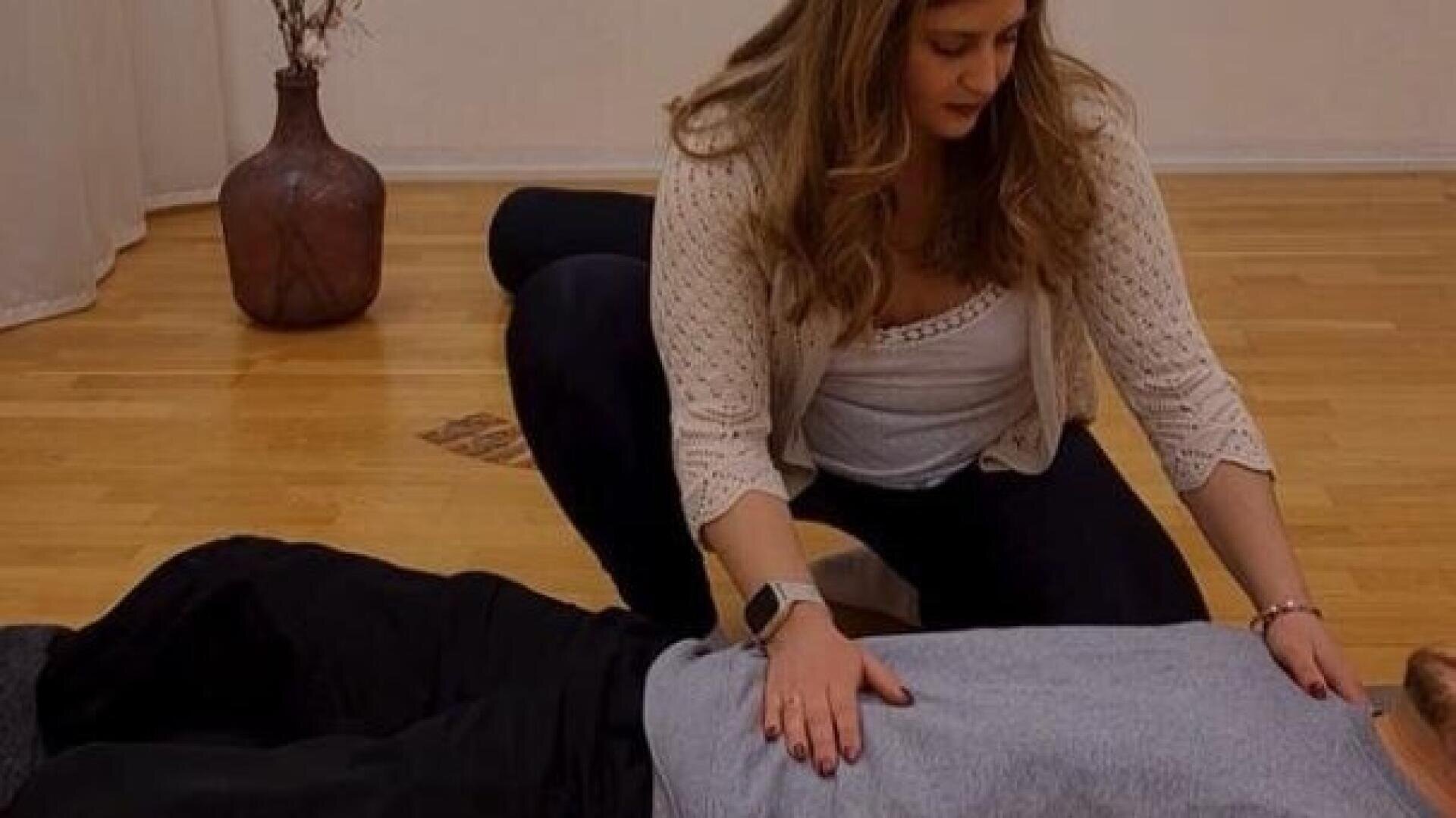 Eine Frau kniet neben einer Person, die auf dem Boden liegt, und legt ihre Hände auf deren Brust und Bauch, als ob sie Hilfe leisten würde, in einem Raum mit Holzfußboden und einer großen Vase im Hintergrund.