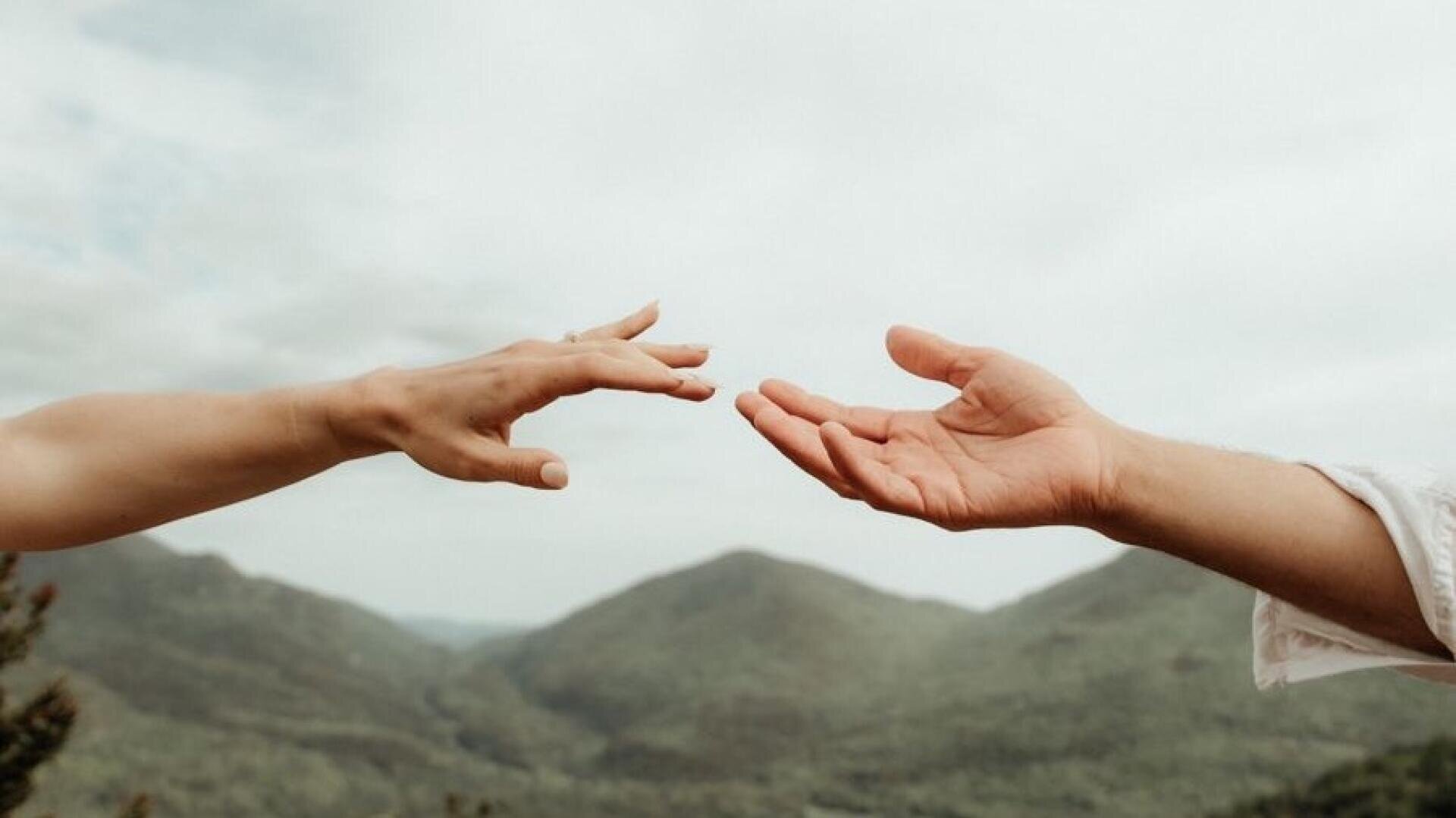 Zwei Hände, die sich entgegenstrecken und deren Finger sich fast berühren, vor dem Hintergrund grüner Berge und eines bewölkten Himmels.