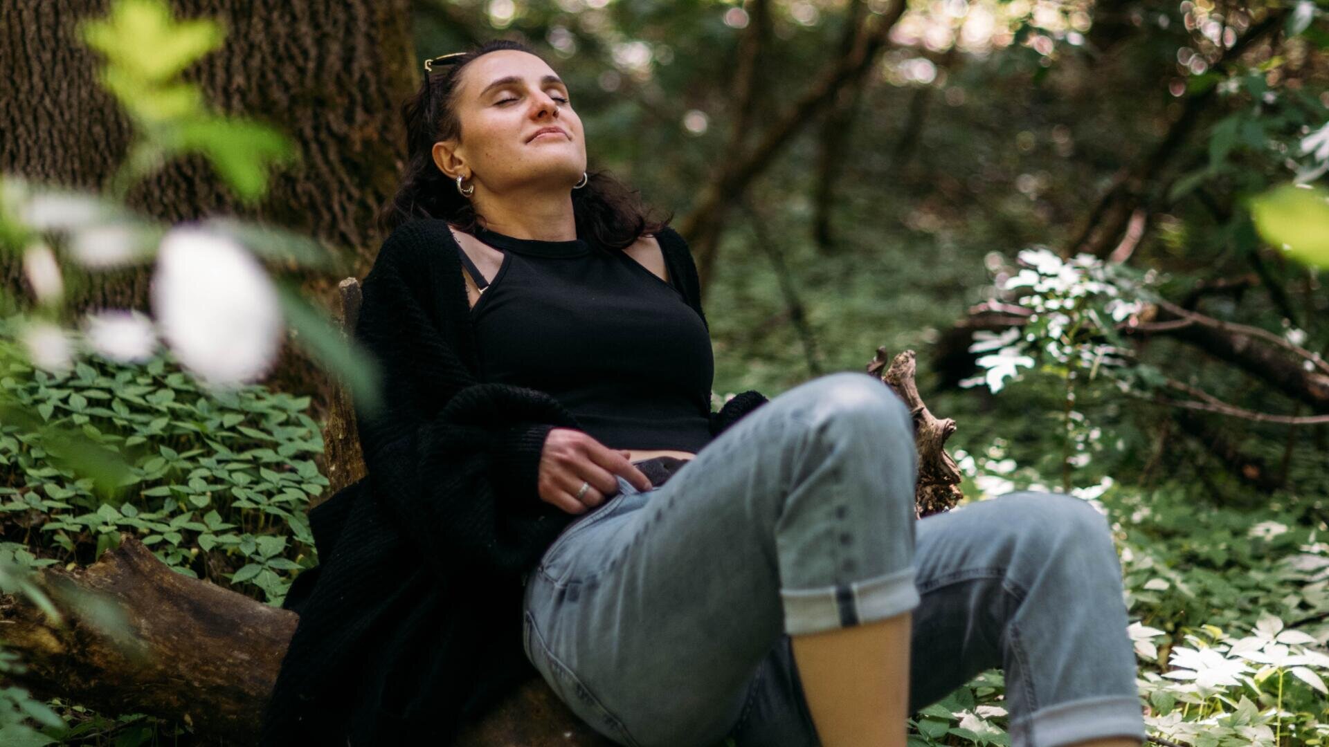 Eine Frau sitzt auf einem umgestürzten Baum in einem Wald, die Augen geschlossen und das Gesicht entspannt, und genießt die friedliche Natur. Sie trägt ein schwarzes Oberteil, Jeans und eine Strickjacke, umgeben von viel Grün.